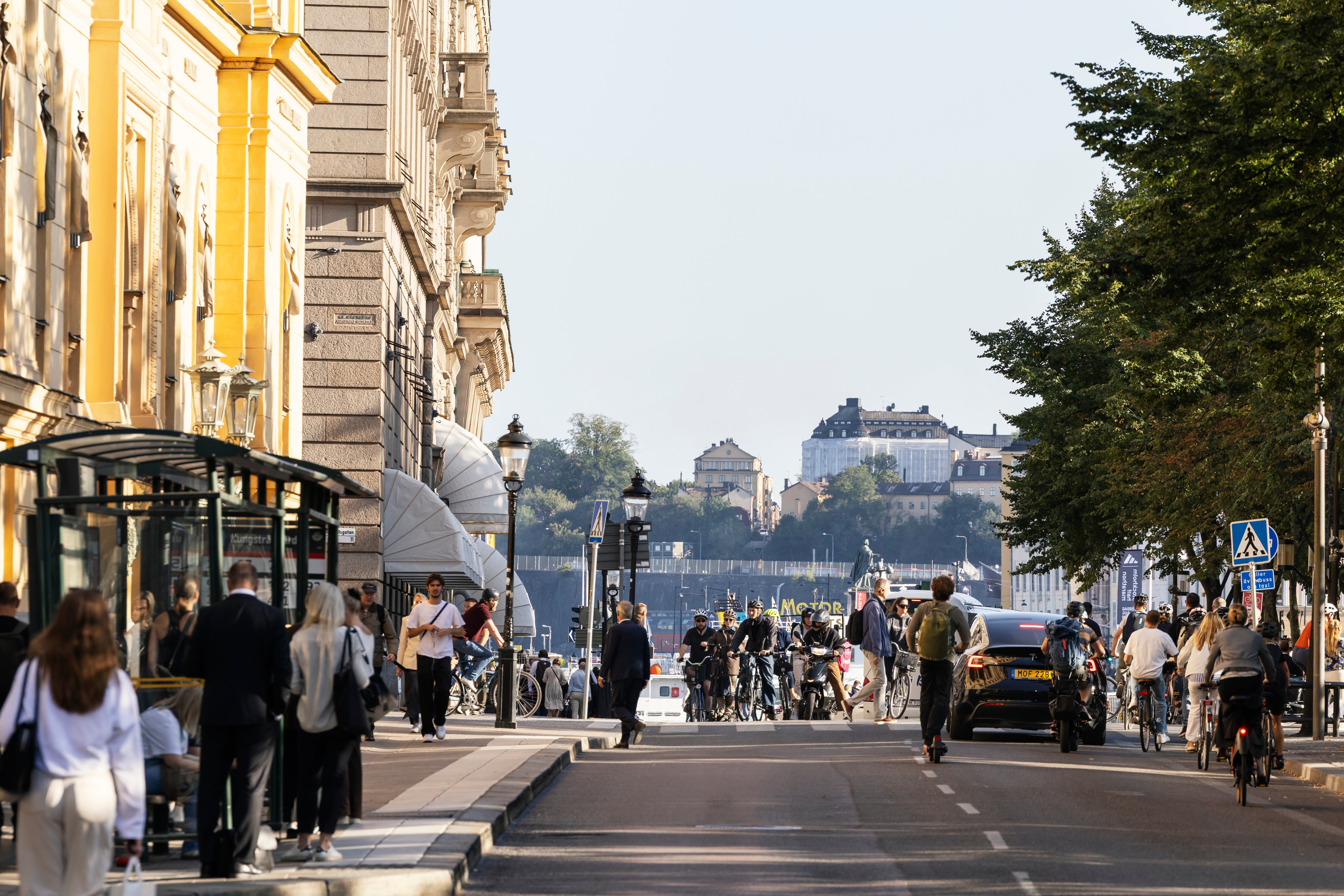 En bred stadsgata med klassiska byggnader på båda sidor. Människor går omkring på gatan. I bakgrunden syns en flod och byggnader på höjderna under en molnig himmel.