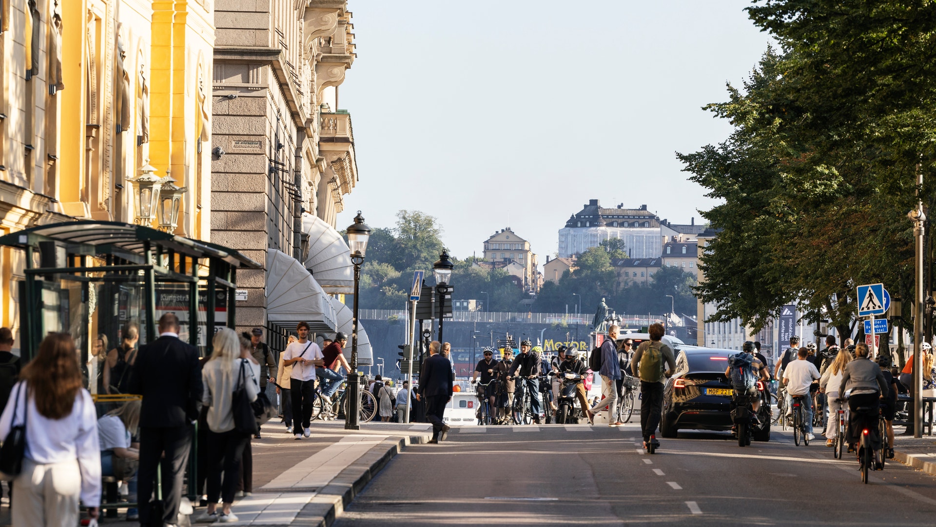 En bred stadsgata med klassiska byggnader på båda sidor. Människor går omkring på gatan. I bakgrunden syns en flod och byggnader på höjderna under en molnig himmel.