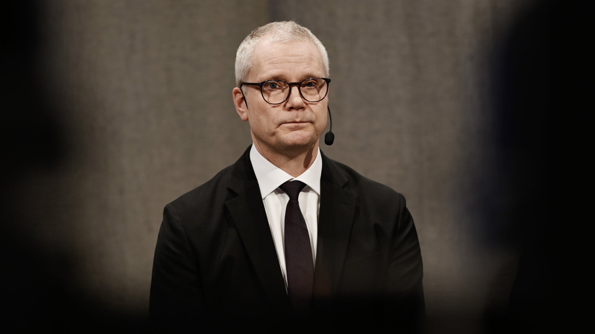 Porträtt av en man i svart kostym och slips med glasögon mot neutral bakgrund.