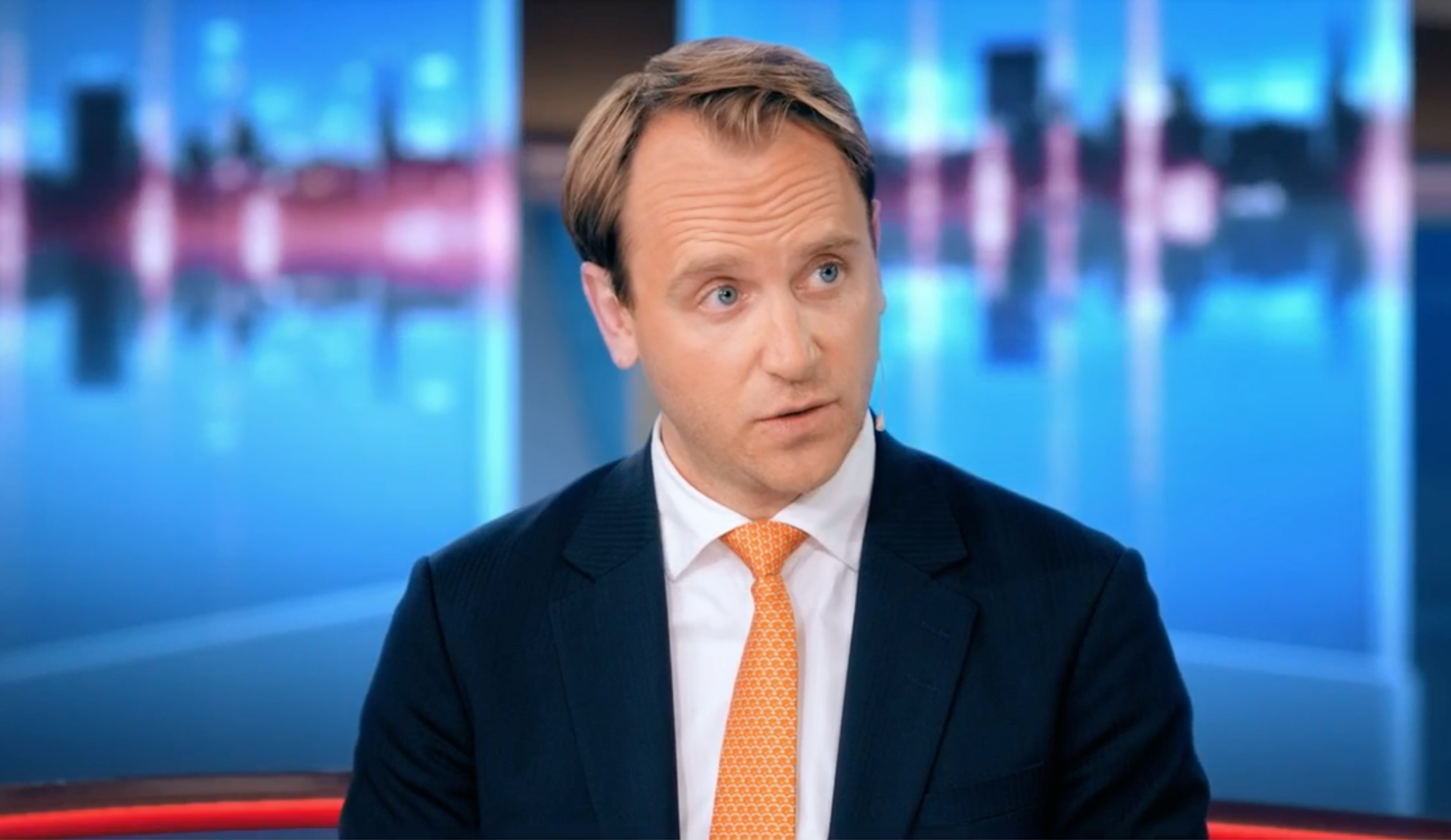 En man i mörk kostym, vit skjorta och orange slips sitter i en tv-studio och tittar åt sidan.