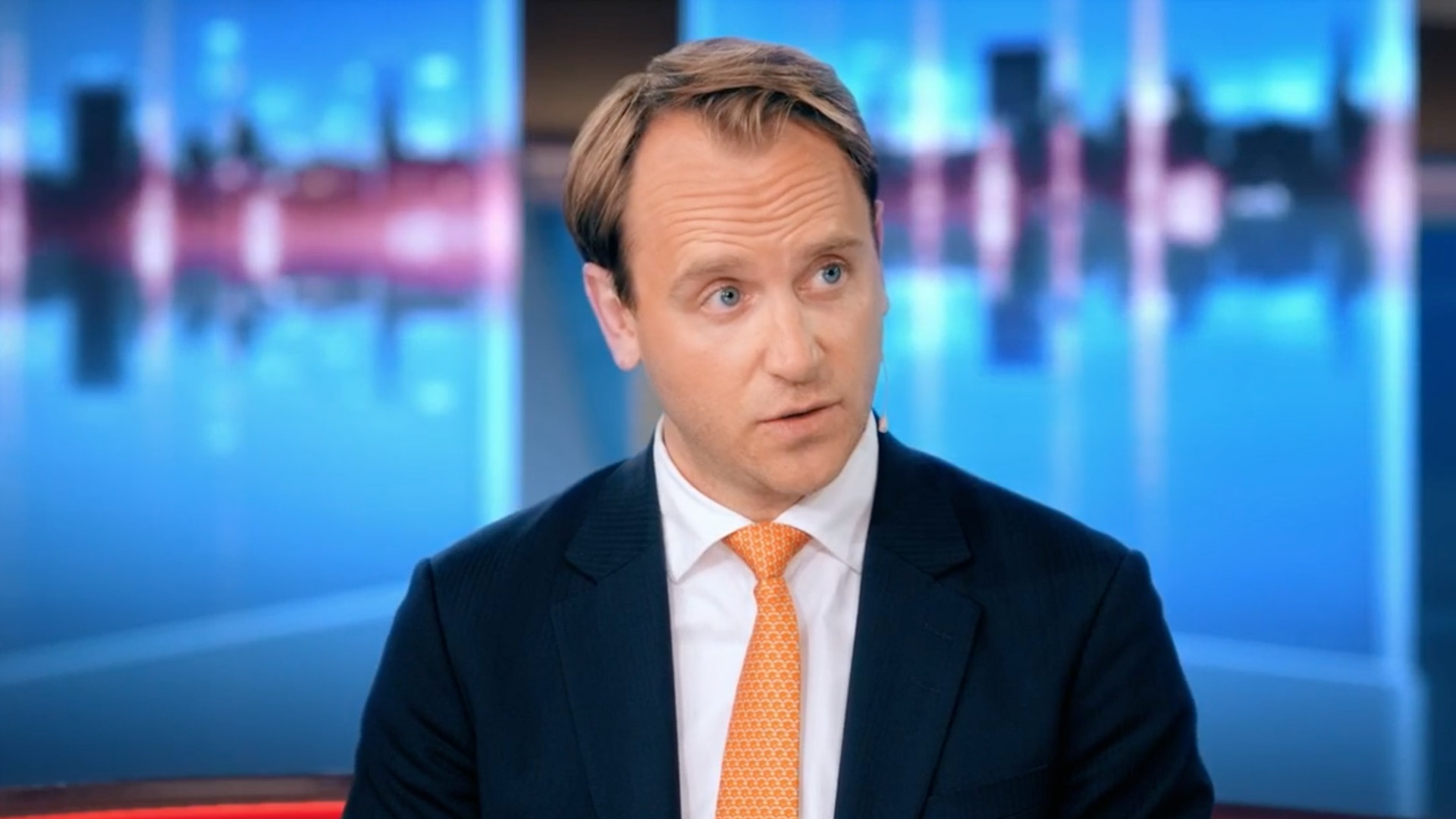 En man i mörk kostym, vit skjorta och orange slips sitter i en tv-studio och tittar åt sidan.