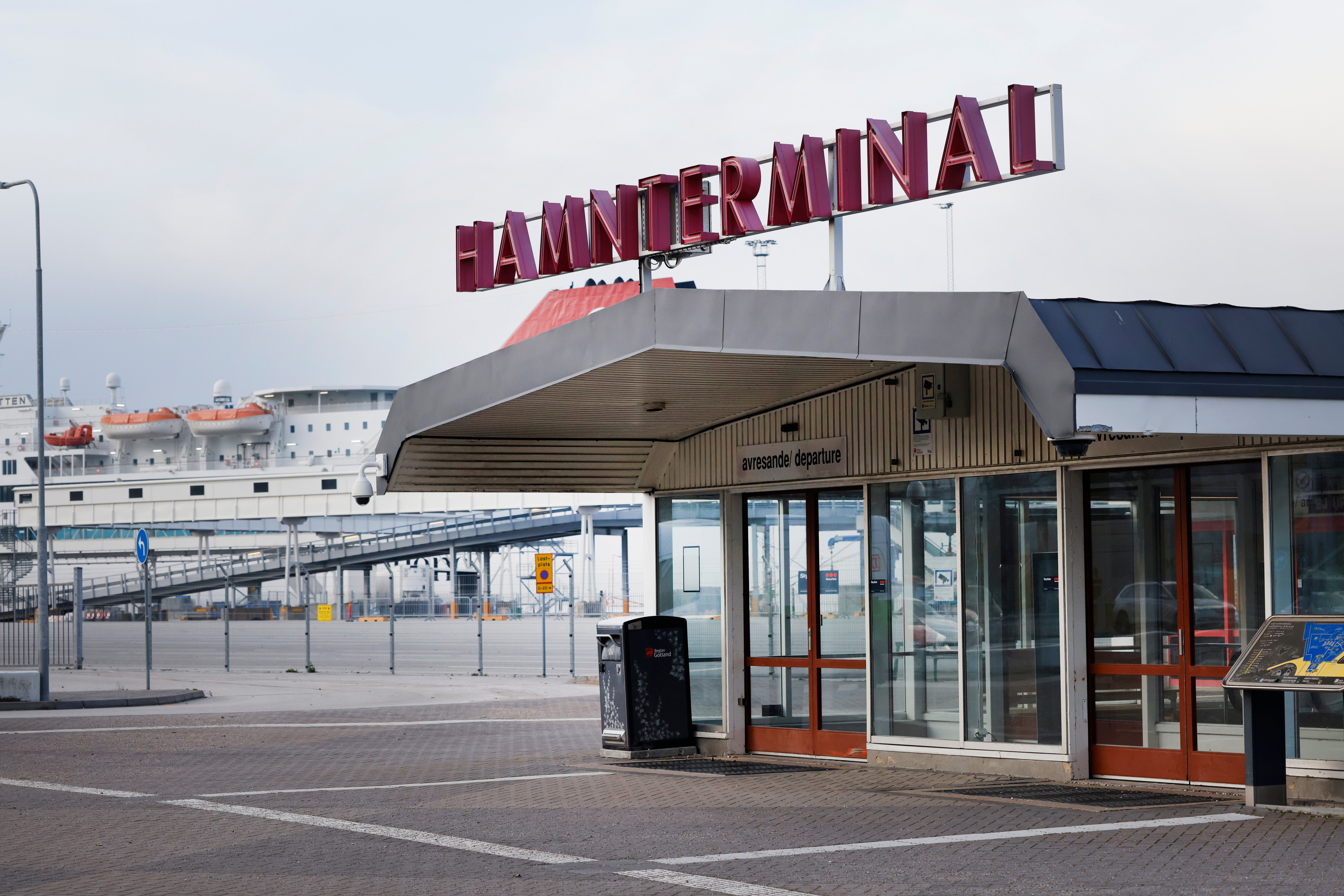 Hamnterminal-byggnad med stor röd skylt vid en hamn, omgiven av metallräcke och moderna byggnader i bakgrunden.