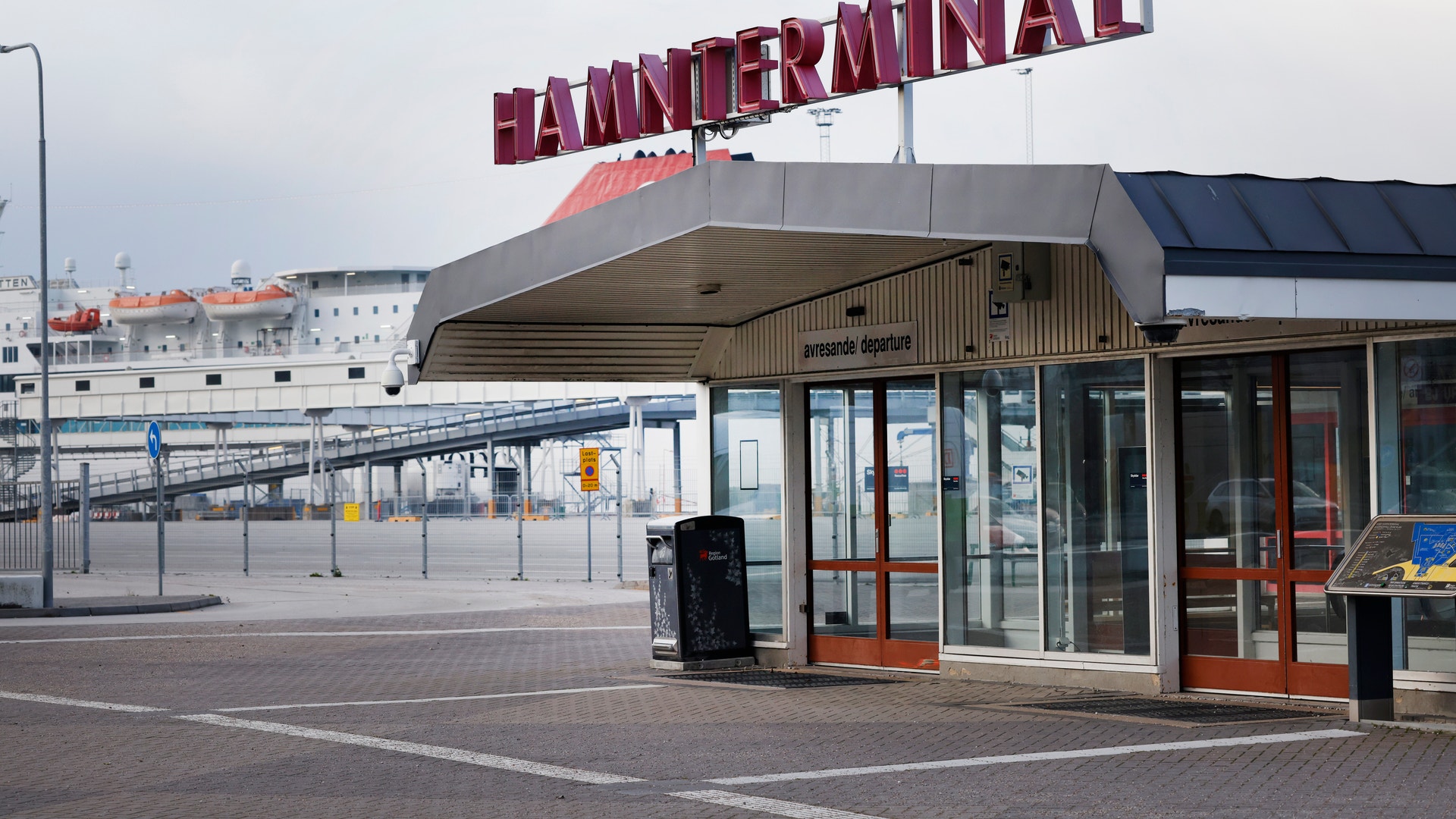 Hamnterminal-byggnad med stor röd skylt vid en hamn, omgiven av metallräcke och moderna byggnader i bakgrunden.