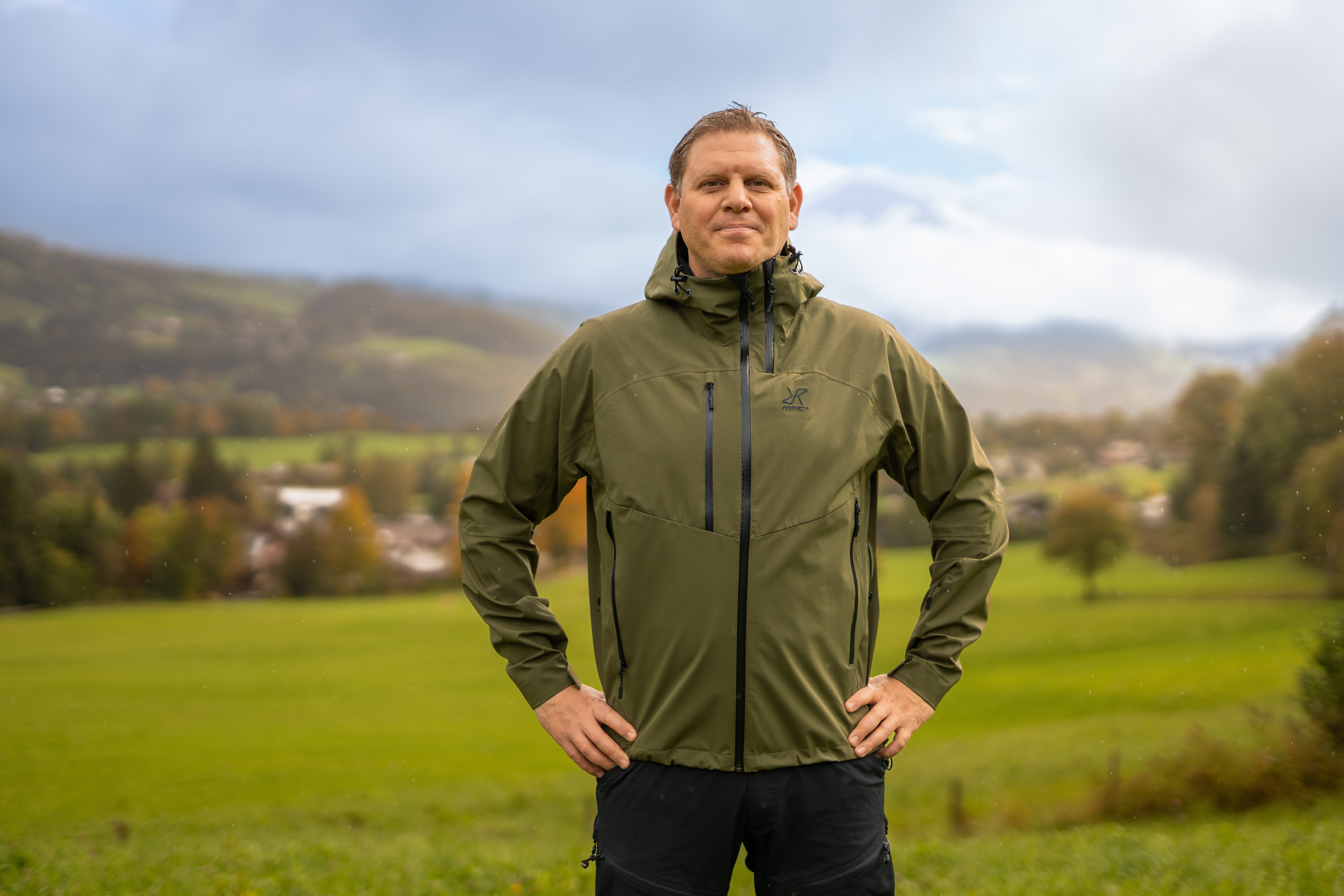 Man i olivgrön jacka står i ett grönt landskap med berg och byggnader i bakgrunden.
