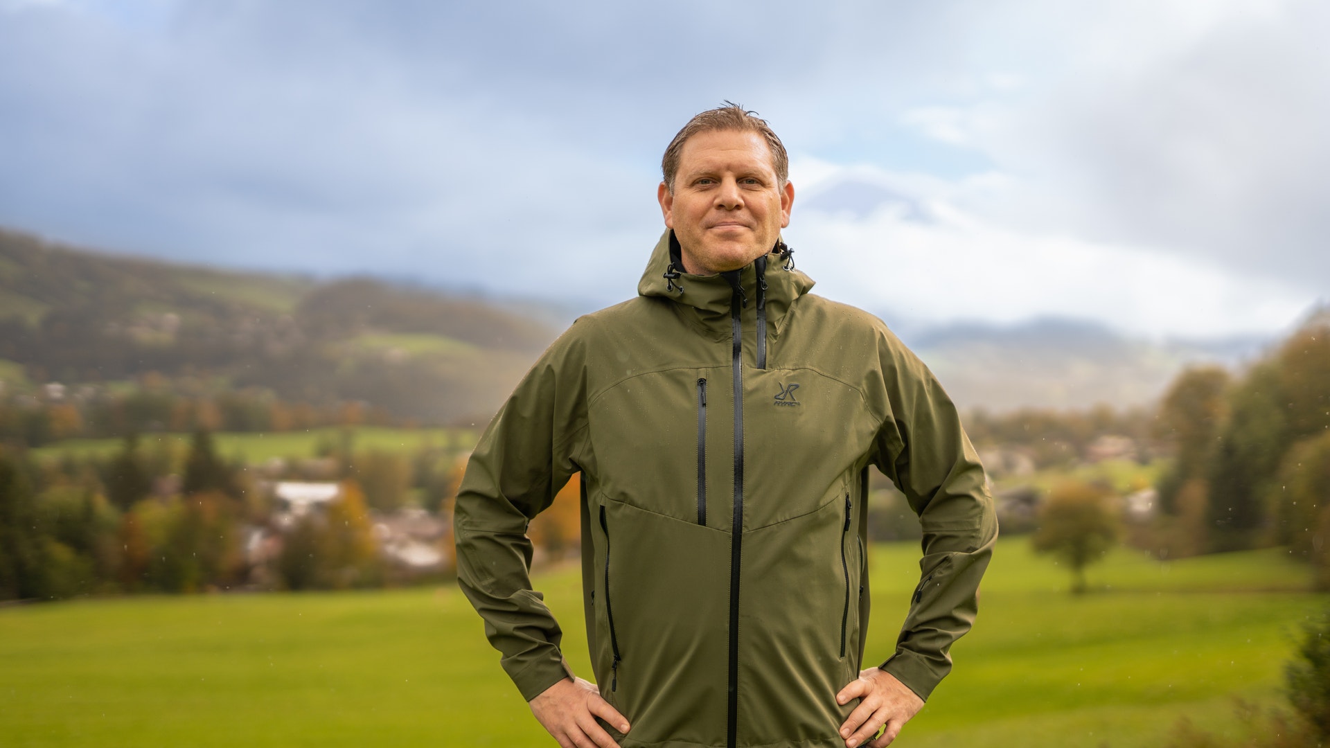 Man i olivgrön jacka står i ett grönt landskap med berg och byggnader i bakgrunden.