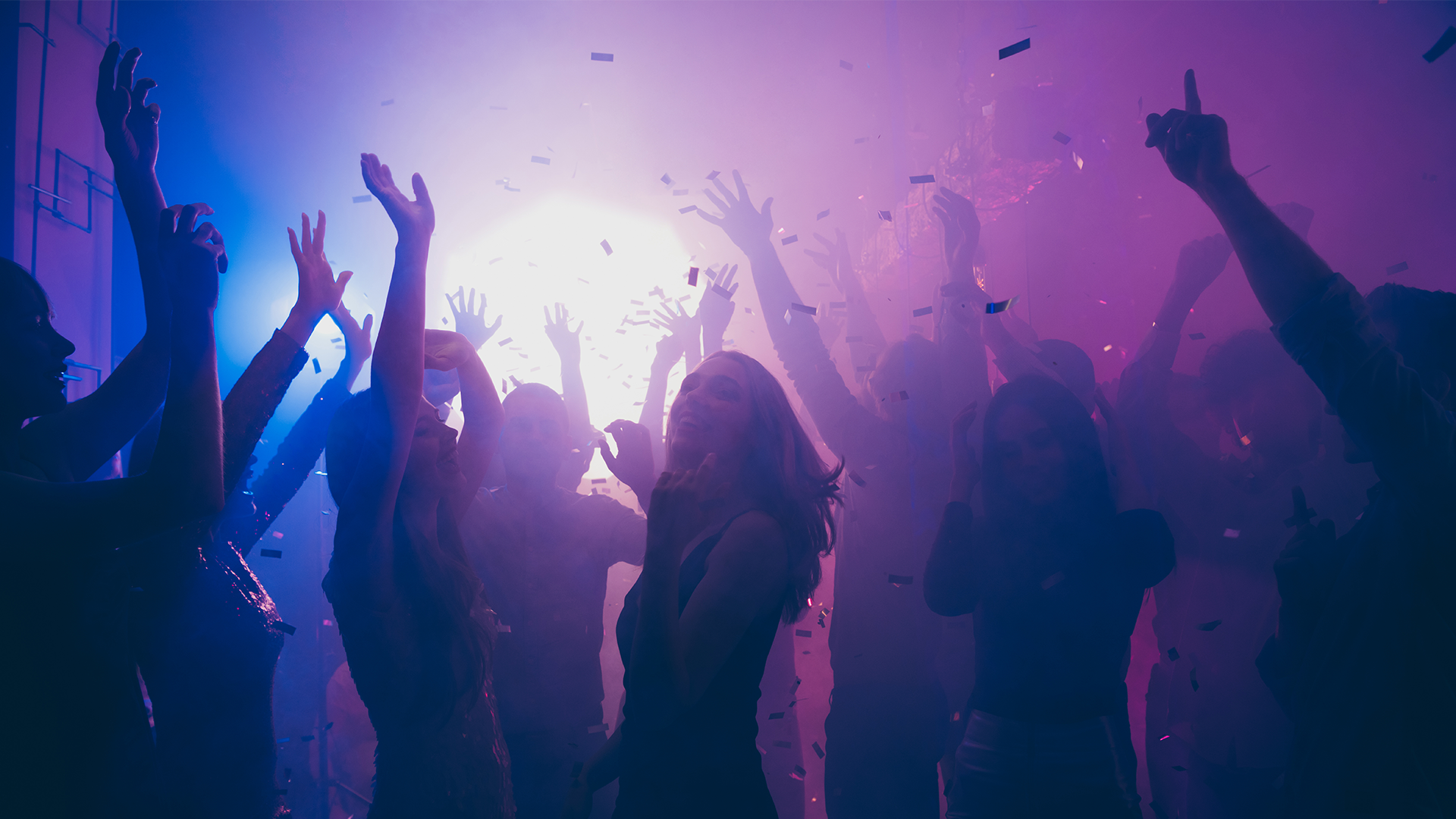 En grupp människor dansar på en fest med händerna upp i luften. Scenen är belyst med lila och blå ljus som skapar en festlig atmosfär.