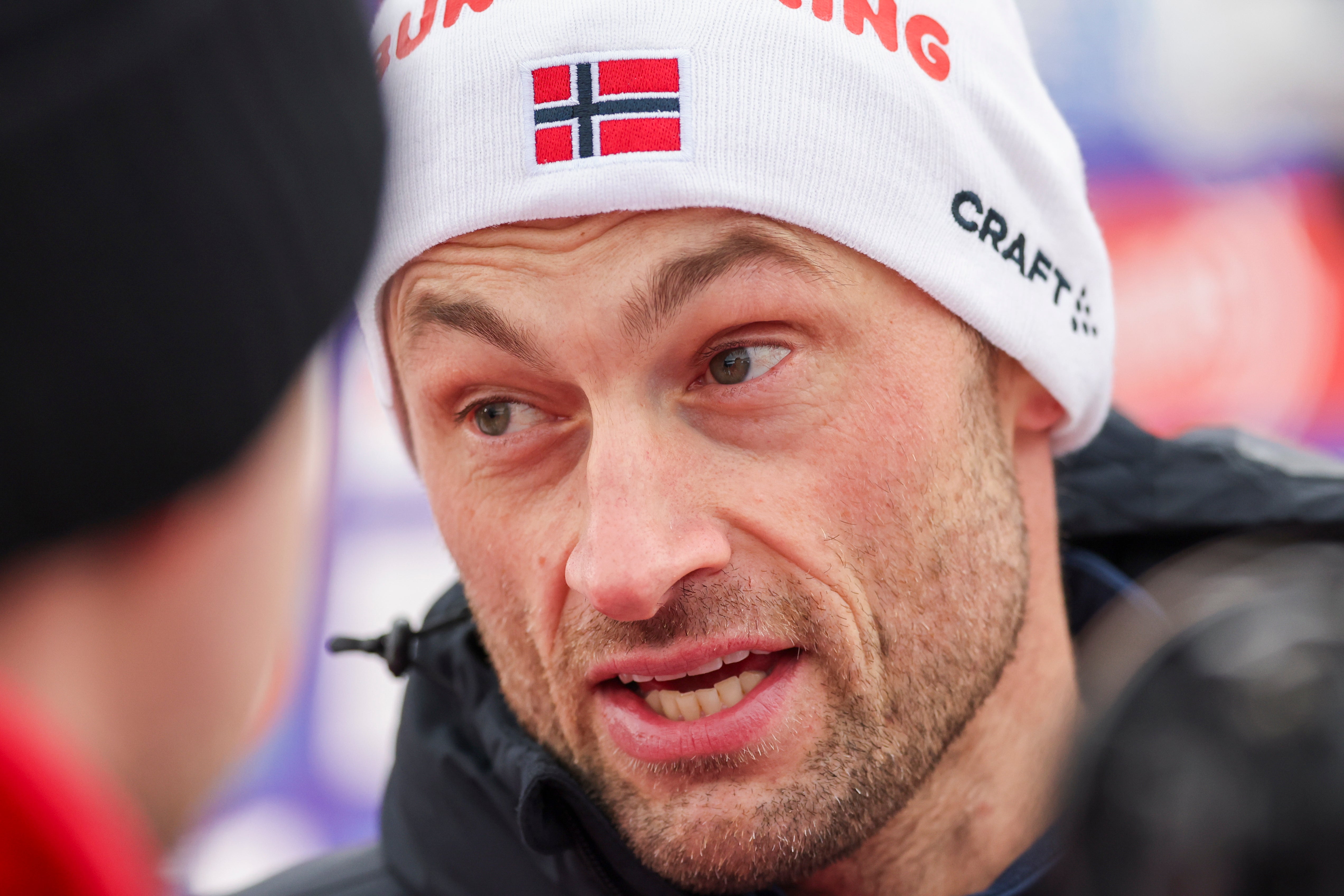 Porträtt av en man med vit mössa med norsk flagga. Han är skidåkare och fotograferad under ett sportevent.