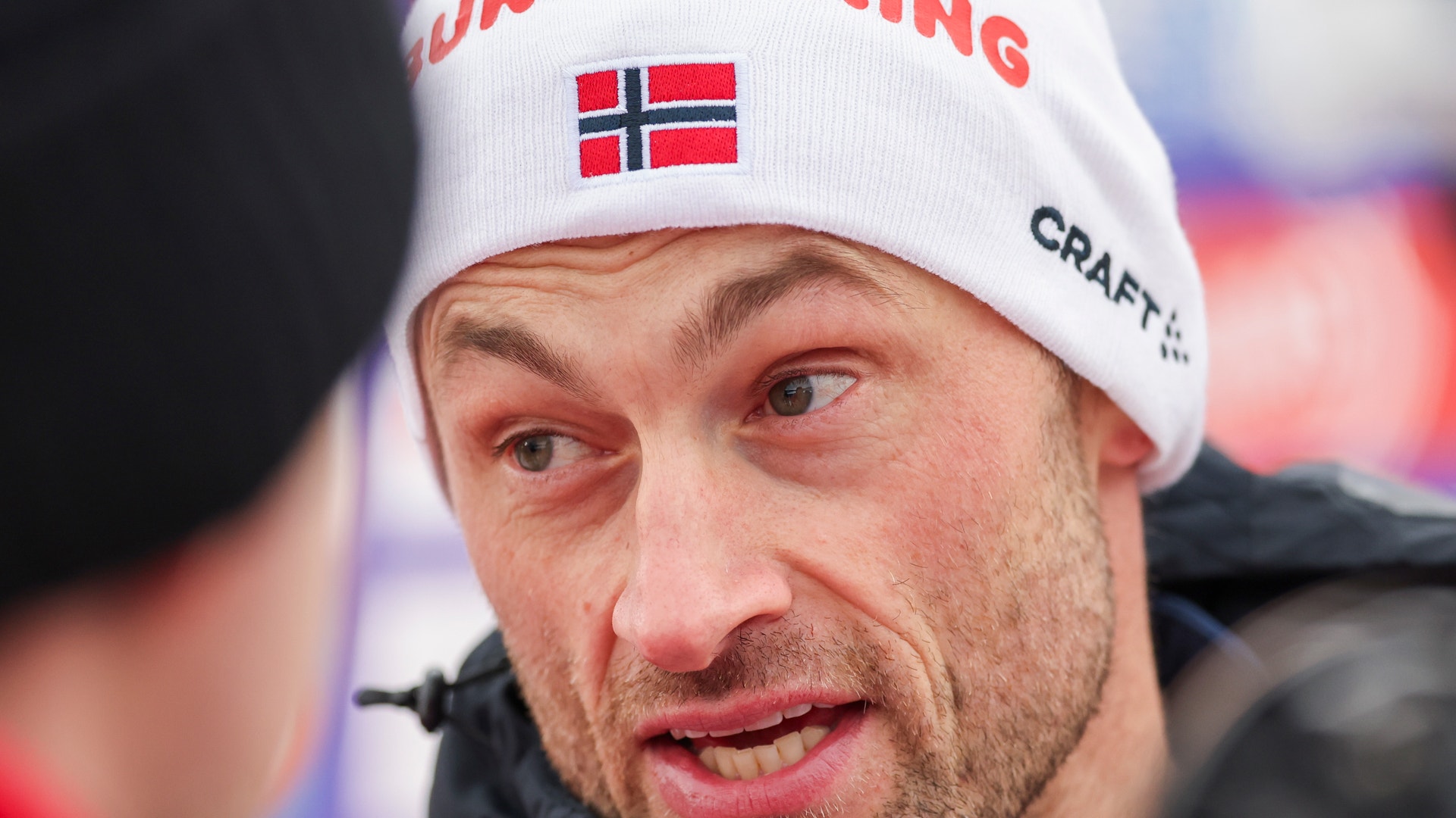 Porträtt av en man med vit mössa med norsk flagga. Han är skidåkare och fotograferad under ett sportevent.