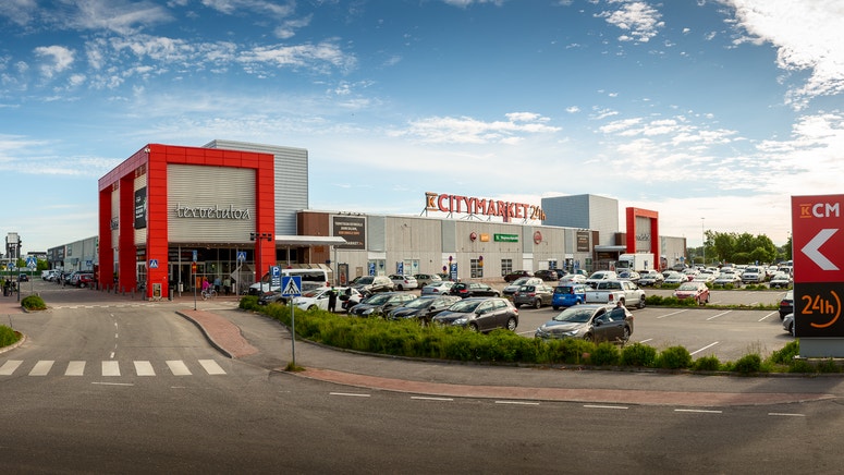 Exterörbild av ett modernt köpcentrum med röda och grå byggnader, parkerade bilar och blå himmel.