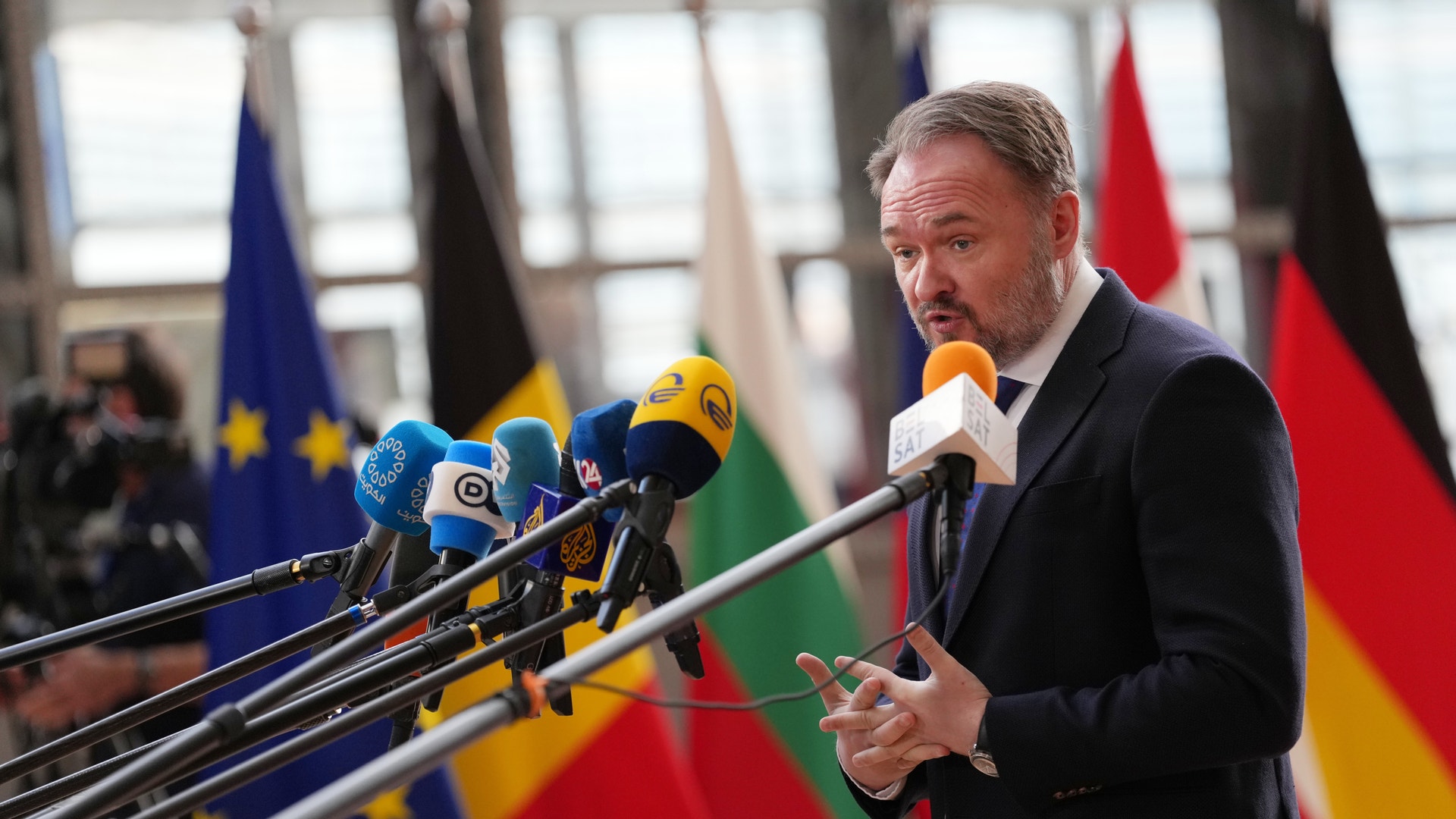 Dan Jørgensen i mörk kostym talar vid ett presskonferens med flera mikrofoner framför honom och internationella flaggor i bakgrunden.
