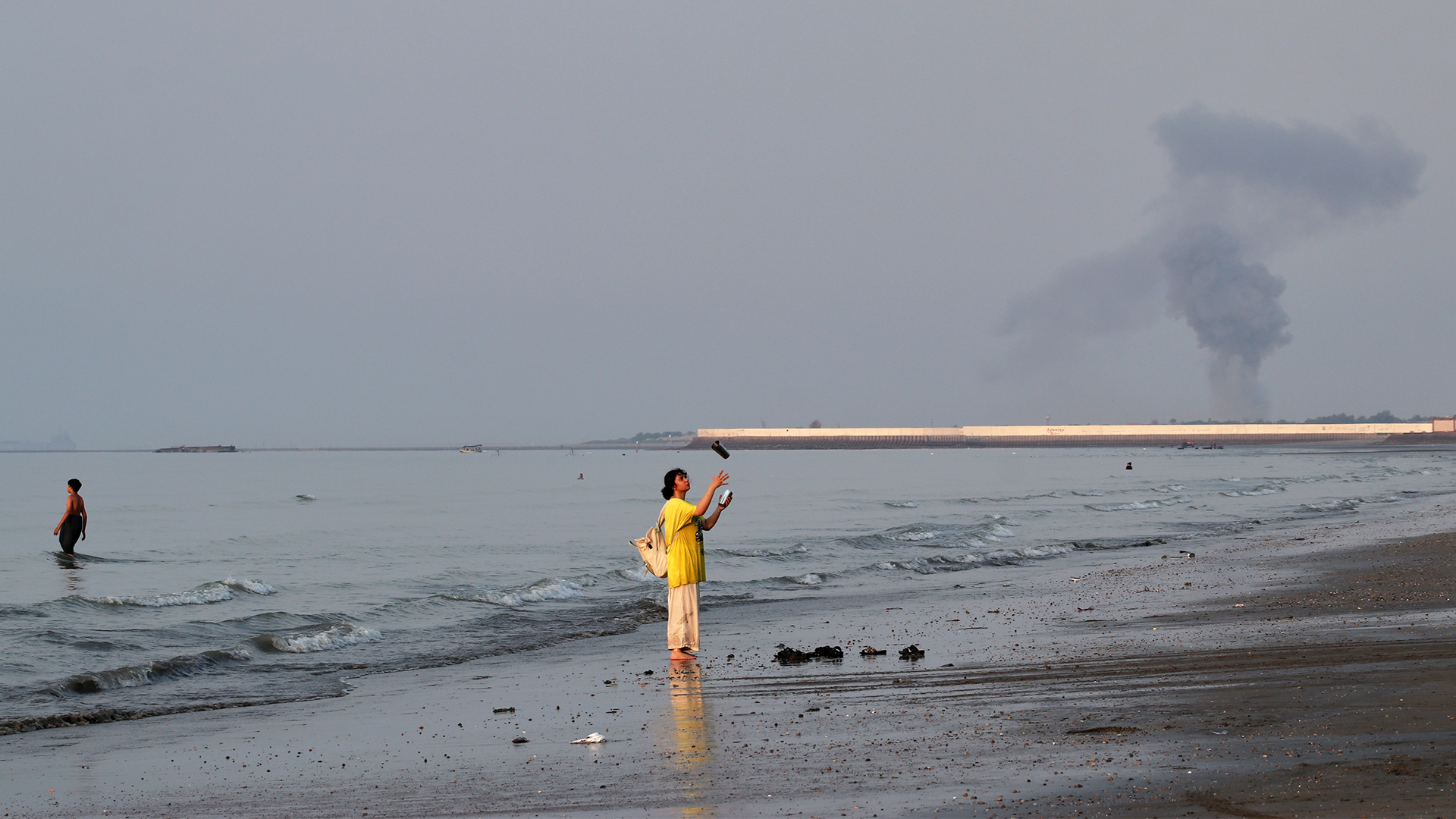 Två personer i vattnet på en strand. En person i gul kläder står i grunt vatten och viftar. En röd och vit pipa sträcker sig över stranden ut i havet. Mulen himmel.