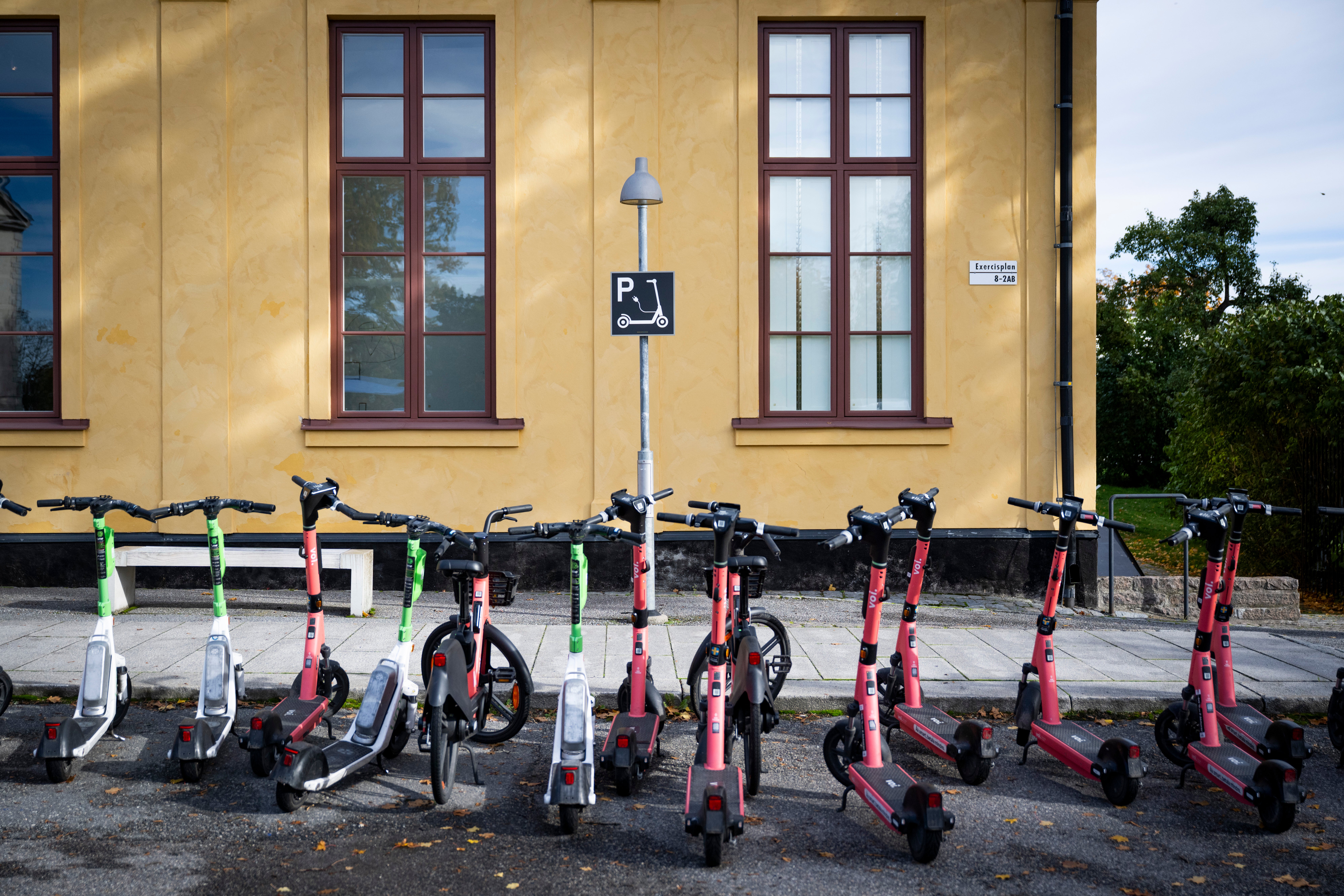 Flera röda elscootrar och gröna cyklar parkerade på rad framför en gul byggnad med bruna fönster och en handikapparkering-skylt.