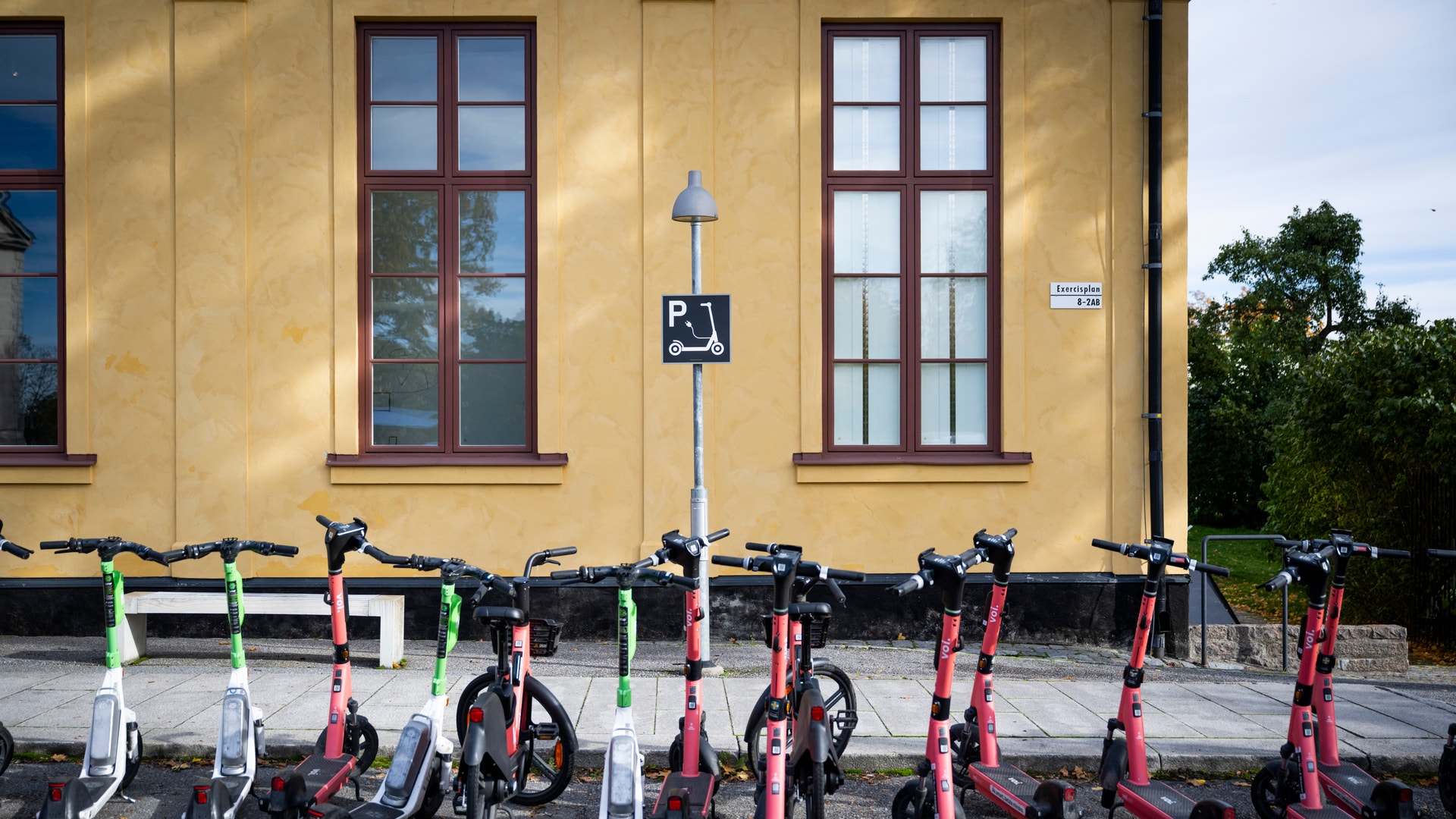 Flera röda elscootrar och gröna cyklar parkerade på rad framför en gul byggnad med bruna fönster och en handikapparkering-skylt.
