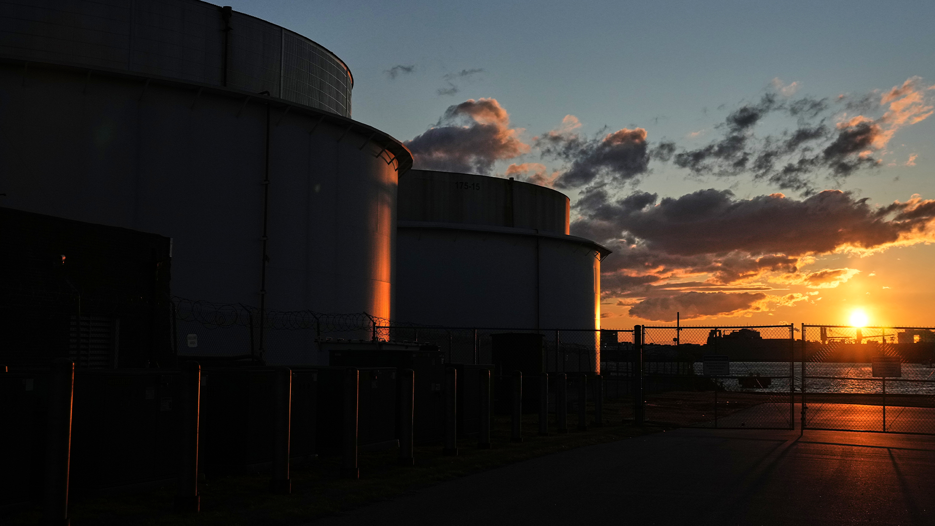 Stora industriella lagertankar silhuetterade mot en dramatisk solnedgång med orange och gula färger på himlen.