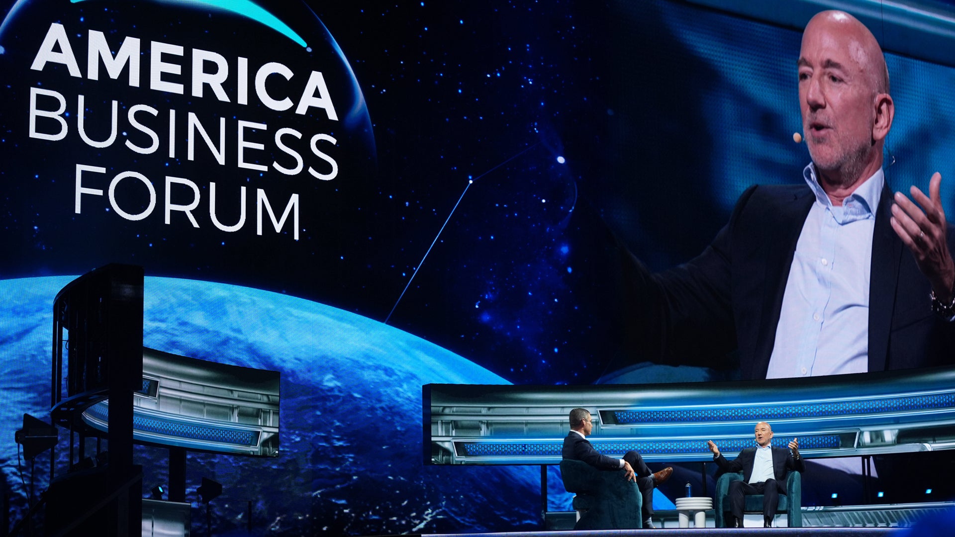 En man presenterar på America Business Forum-konferensen med en stor skärm i bakgrunden och publik i förgrunden.