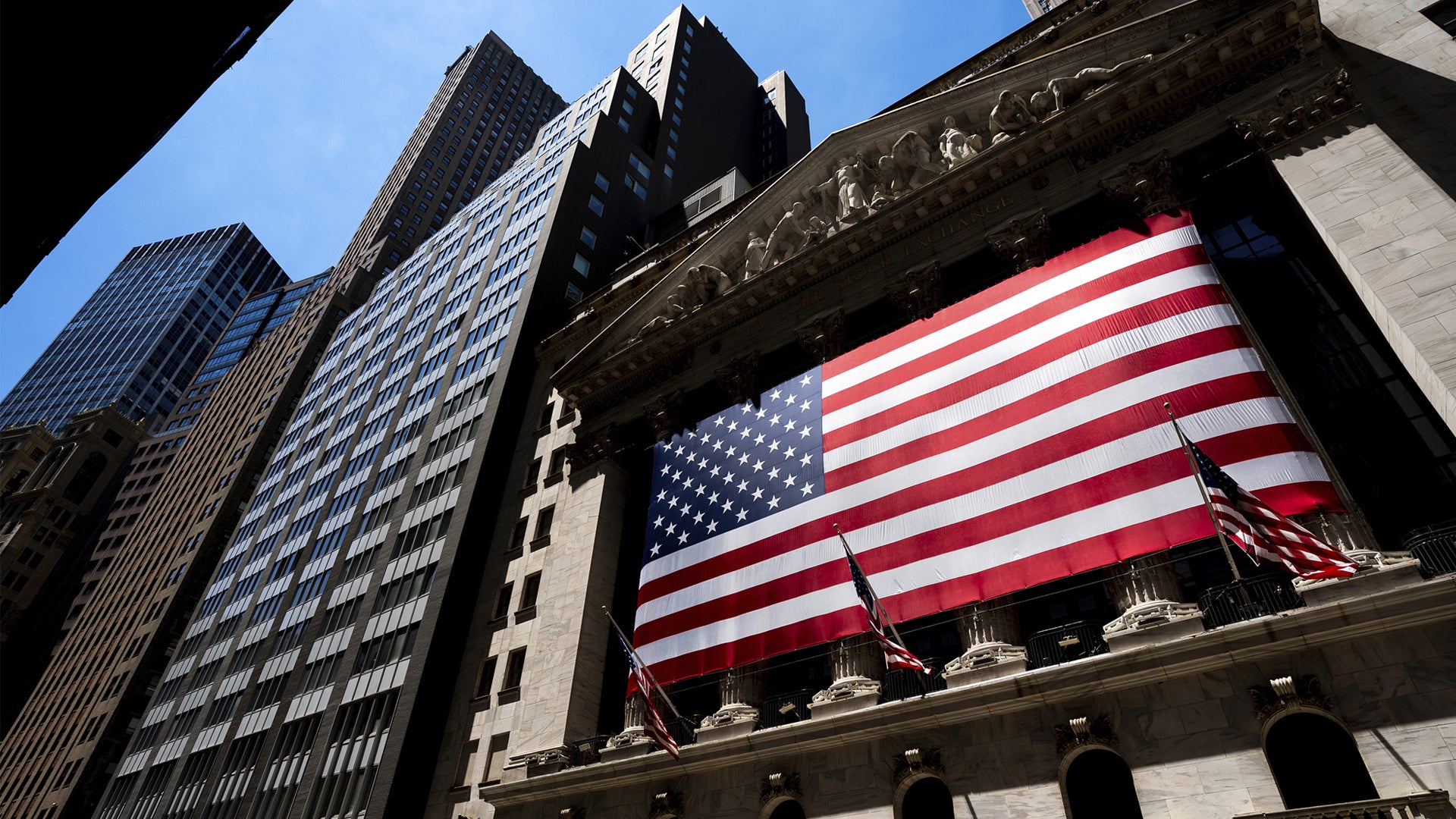 En stor amerikansk flagga hänger på fasaden av New York Stock Exchange, omgiven av höga kontorsbyggnader.