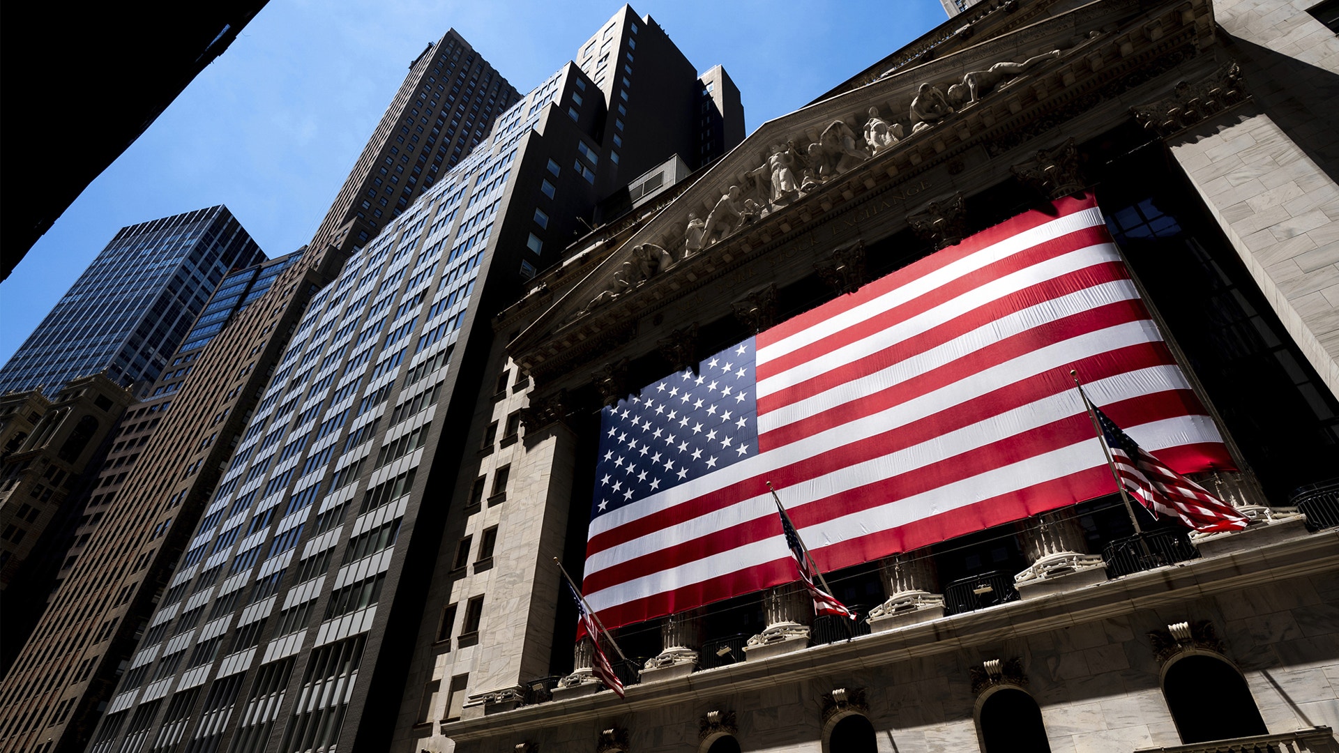 En stor amerikansk flagga hänger på fasaden av New York Stock Exchange, omgiven av höga kontorsbyggnader.