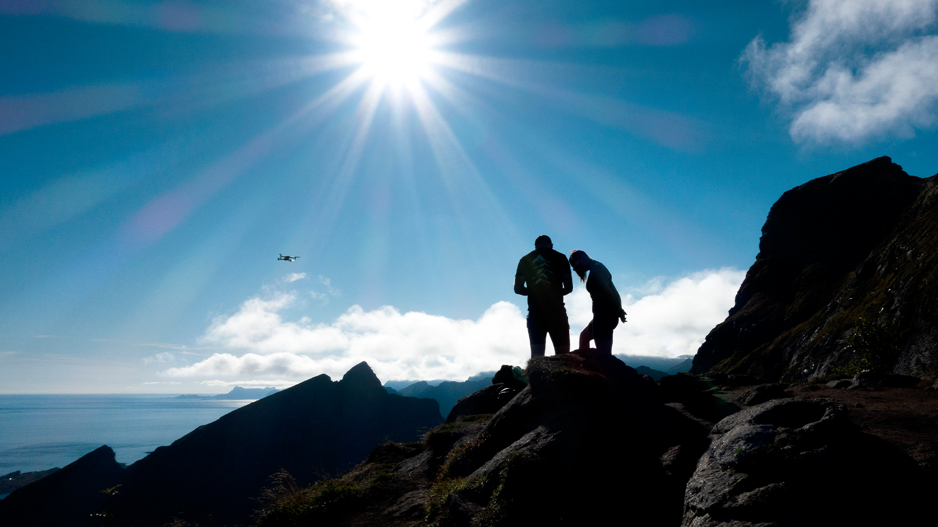 Två personer står på en bergtopp i silhuett mot en ljus himmel med sol och moln. Havet syns i bakgrunden.