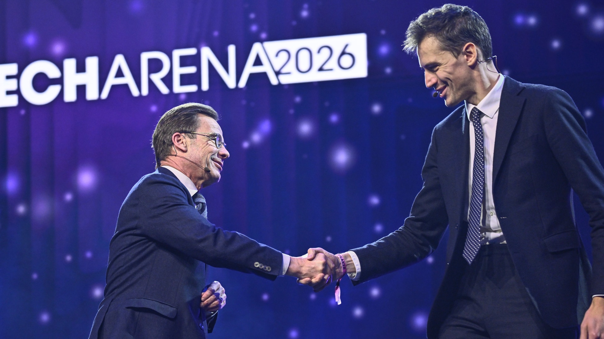 Två män i mörka kostymer skakar hand på en scen under ECHARENA 2026-evenemanget. Bakgrunden är mörkblå med ljuseffekter.