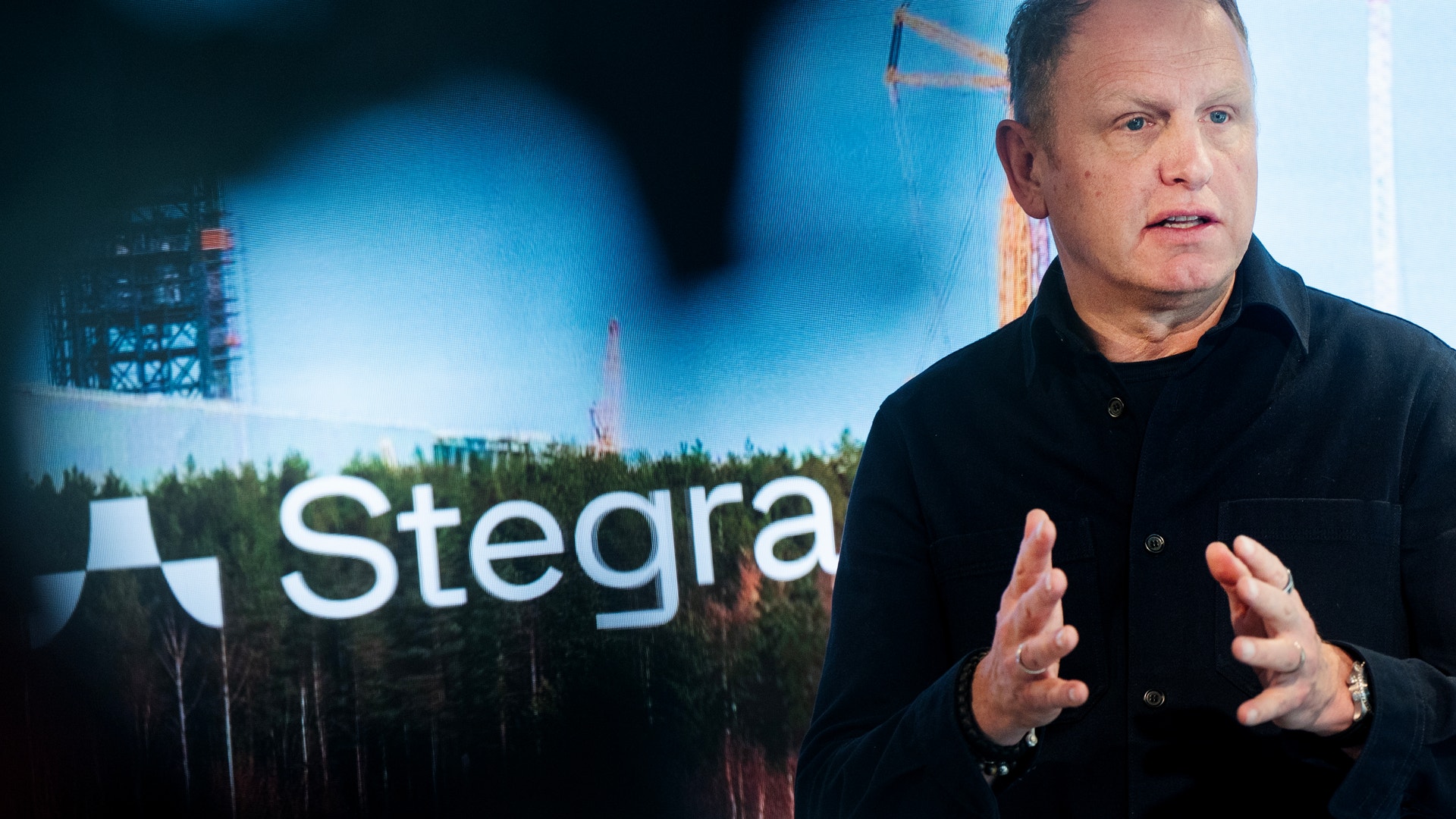 Man i svart skjorta presenterar framför Stegra-logotyp och industriell bakgrund.