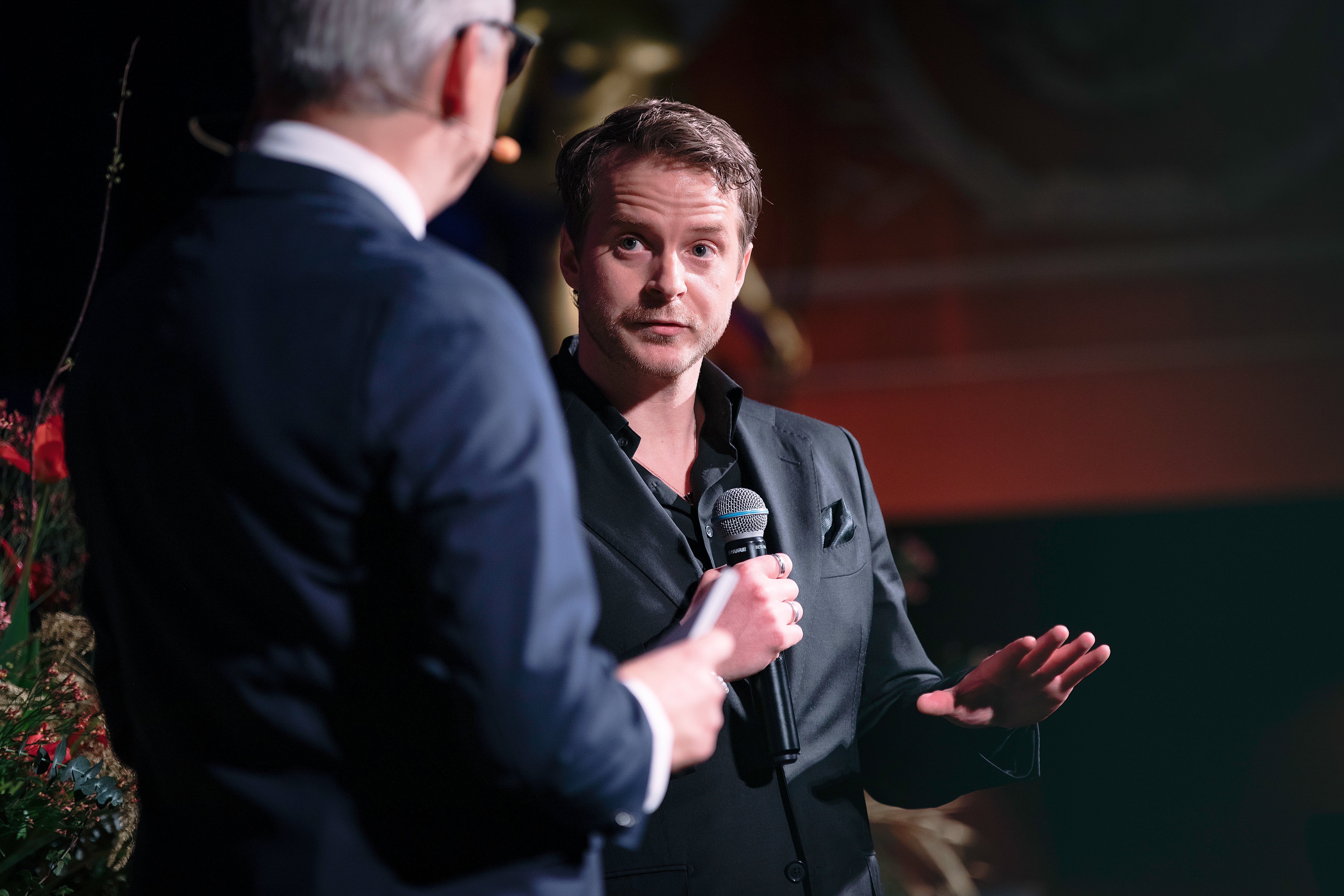 Två män i samtal på en scen. Mannen till höger håller i en mikrofon och verkar tala eller bli intervjuad.