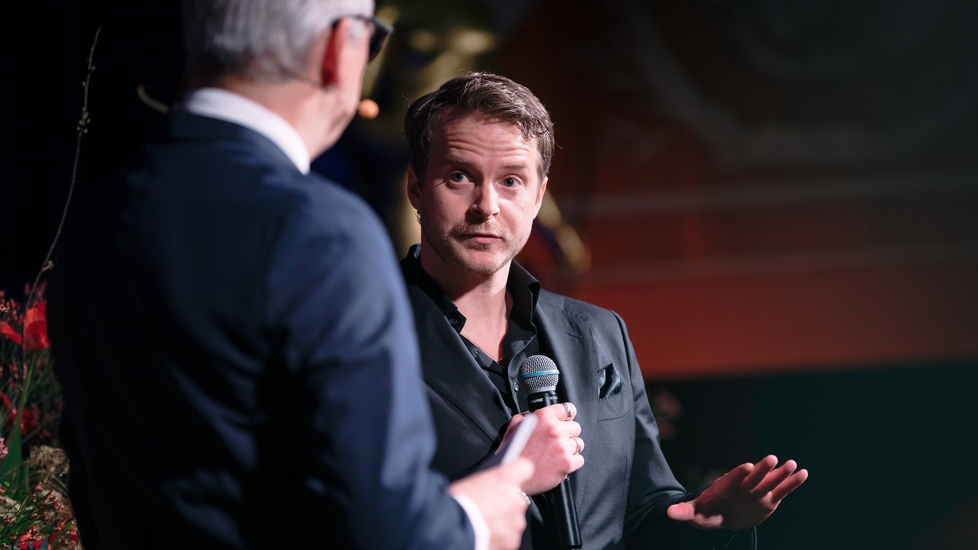 Två män i samtal på en scen. Mannen till höger håller i en mikrofon och verkar tala eller bli intervjuad.