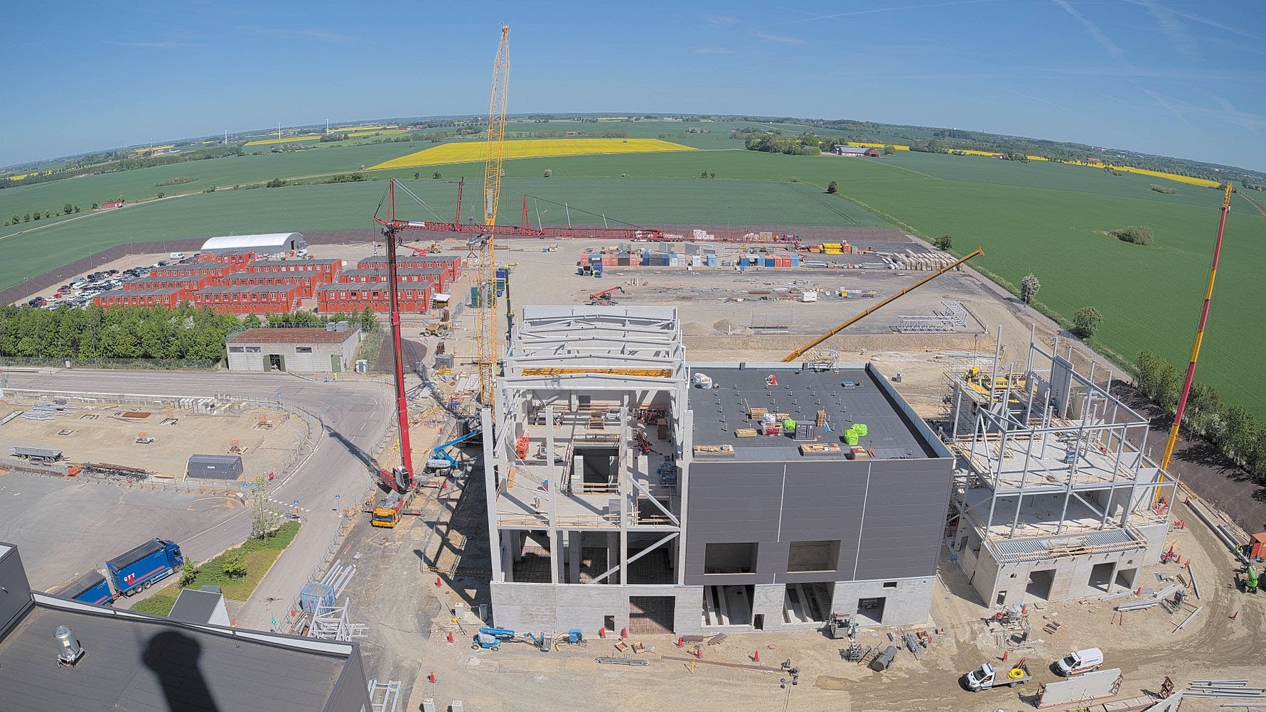 Flygfoto av en byggarbetsplats med en industribyggnad under konstruktion. Betongstrukturer, kranar och byggnadsställningar syns på platsen omgiven av gröna åkrar.