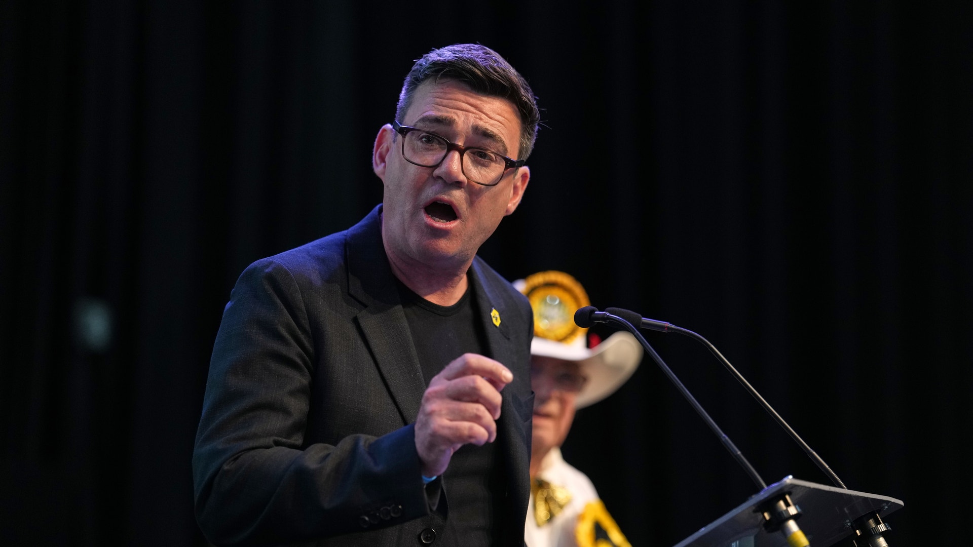 En man med glasögon talar vid en mikrofon på en scen. Han bär en mörk blazer och gestikulerar medan han presenterar. En guldpokal syns i bakgrunden.