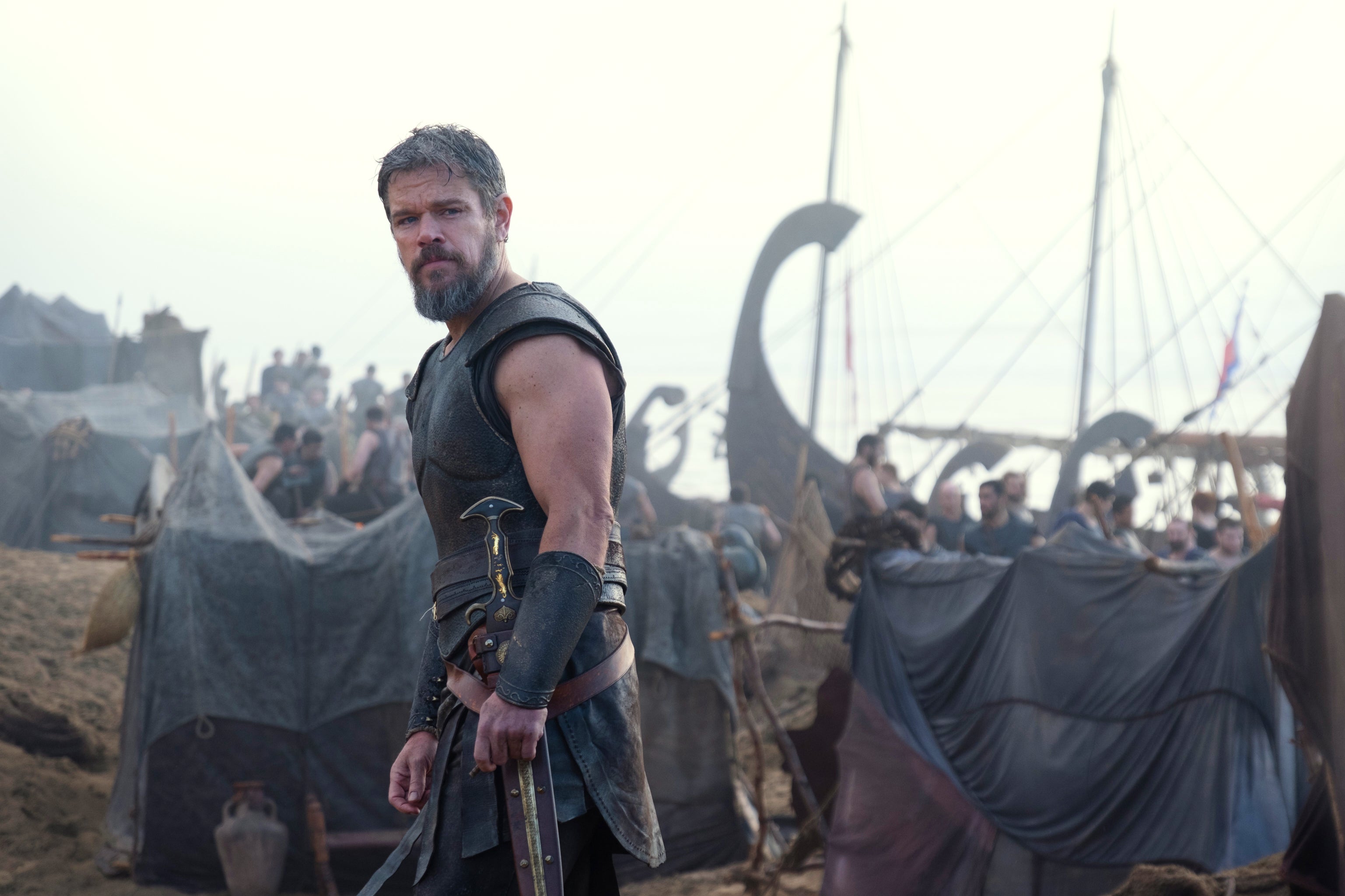 Man i medeltida rustning och rustade kläder står framför ett stort vikingalager med båtar och många människor i bakgrunden.