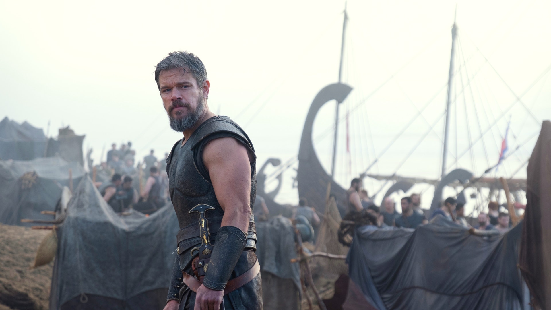 Man i medeltida rustning och rustade kläder står framför ett stort vikingalager med båtar och många människor i bakgrunden.