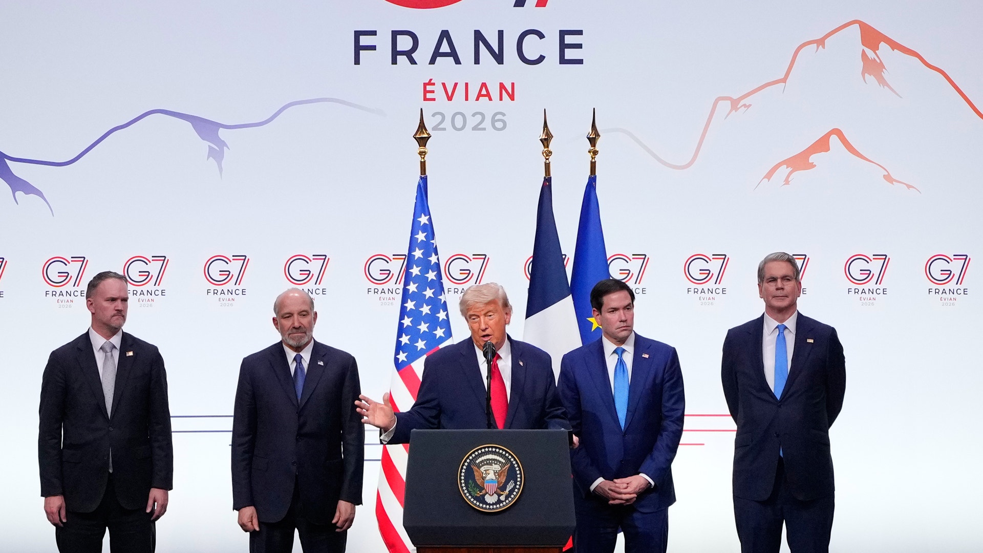 En amerikansk president talar vid ett podium under ett G7-möte i Frankrike. Han är omgiven av fyra män i kostymer, med amerikanska och franska flaggor i bakgrunden.