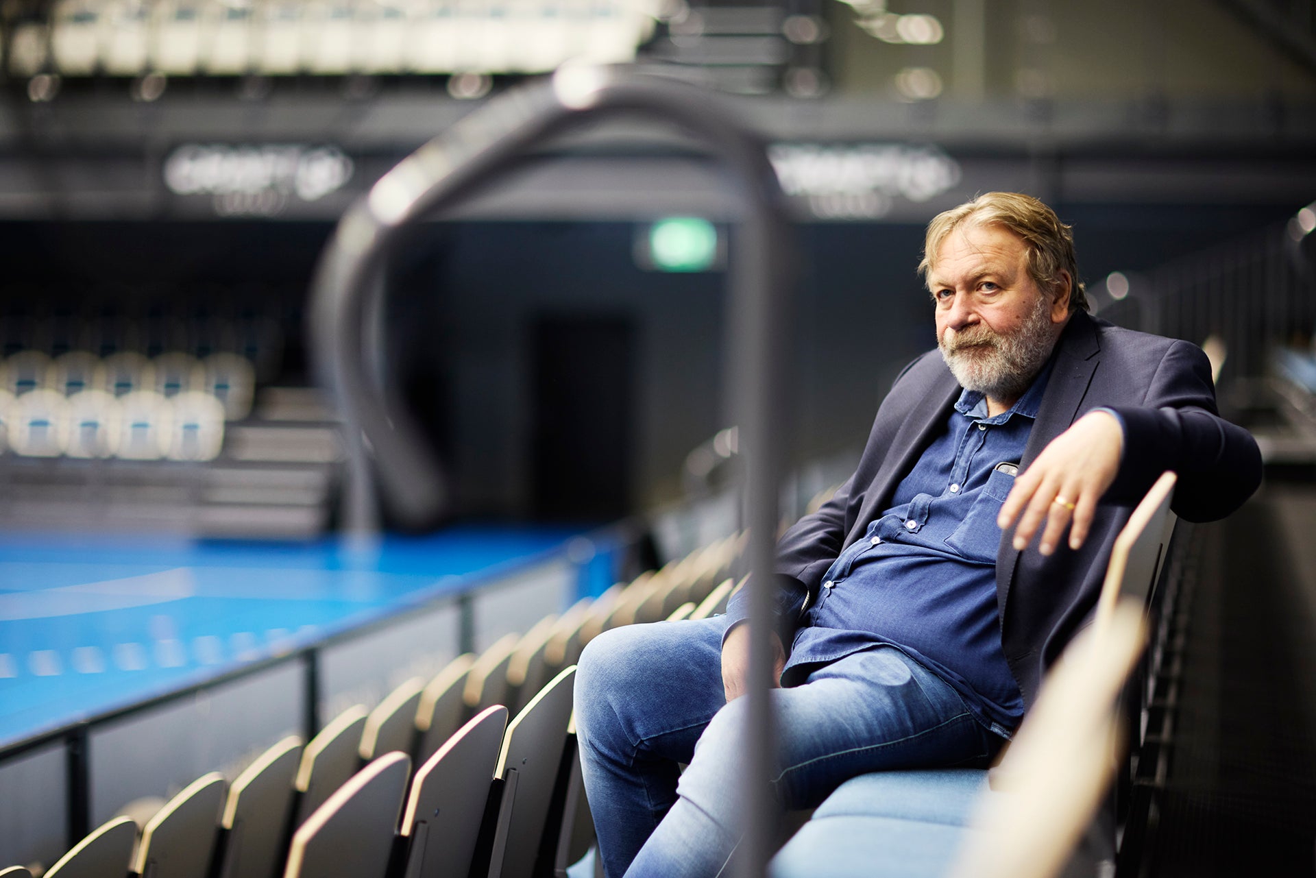 En man med grått hår och skägg sitter på läktaren i en stor idrottshall. Han bär en mörkblå skjorta och ser avslappnad ut. I bakgrunden syns en blå idrottsplan.