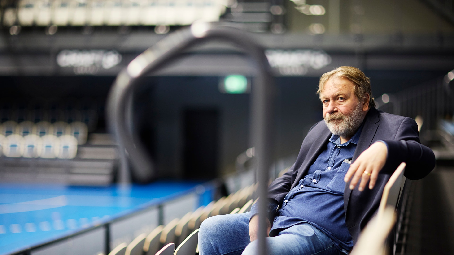 En man med grått hår och skägg sitter på läktaren i en stor idrottshall. Han bär en mörkblå skjorta och ser avslappnad ut. I bakgrunden syns en blå idrottsplan.