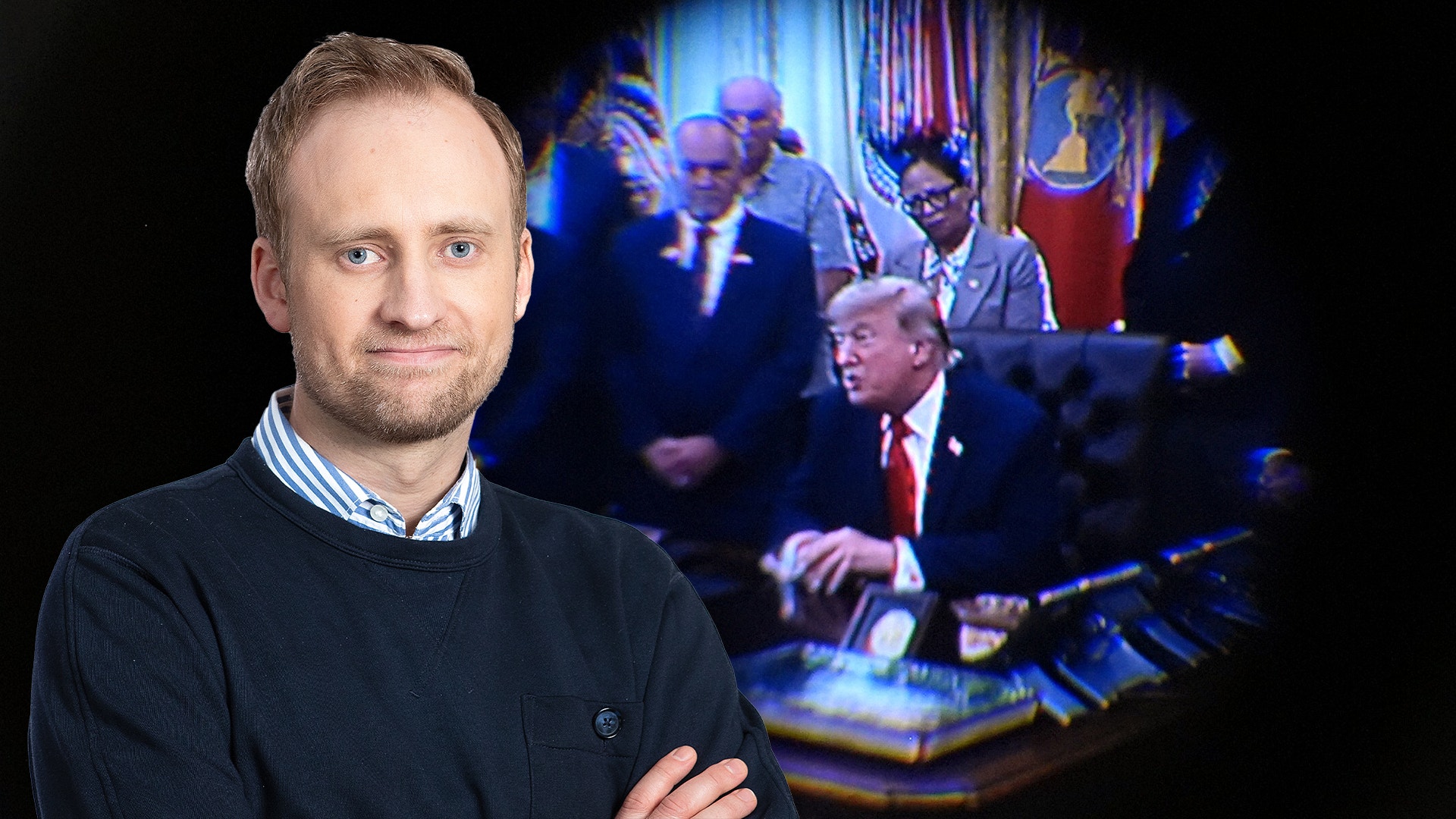 En man i mörk tröja står i förgrunden med armarna i kors. I bakgrunden syns en suddig scen från ett officiellt möte med flera personer vid ett bord.