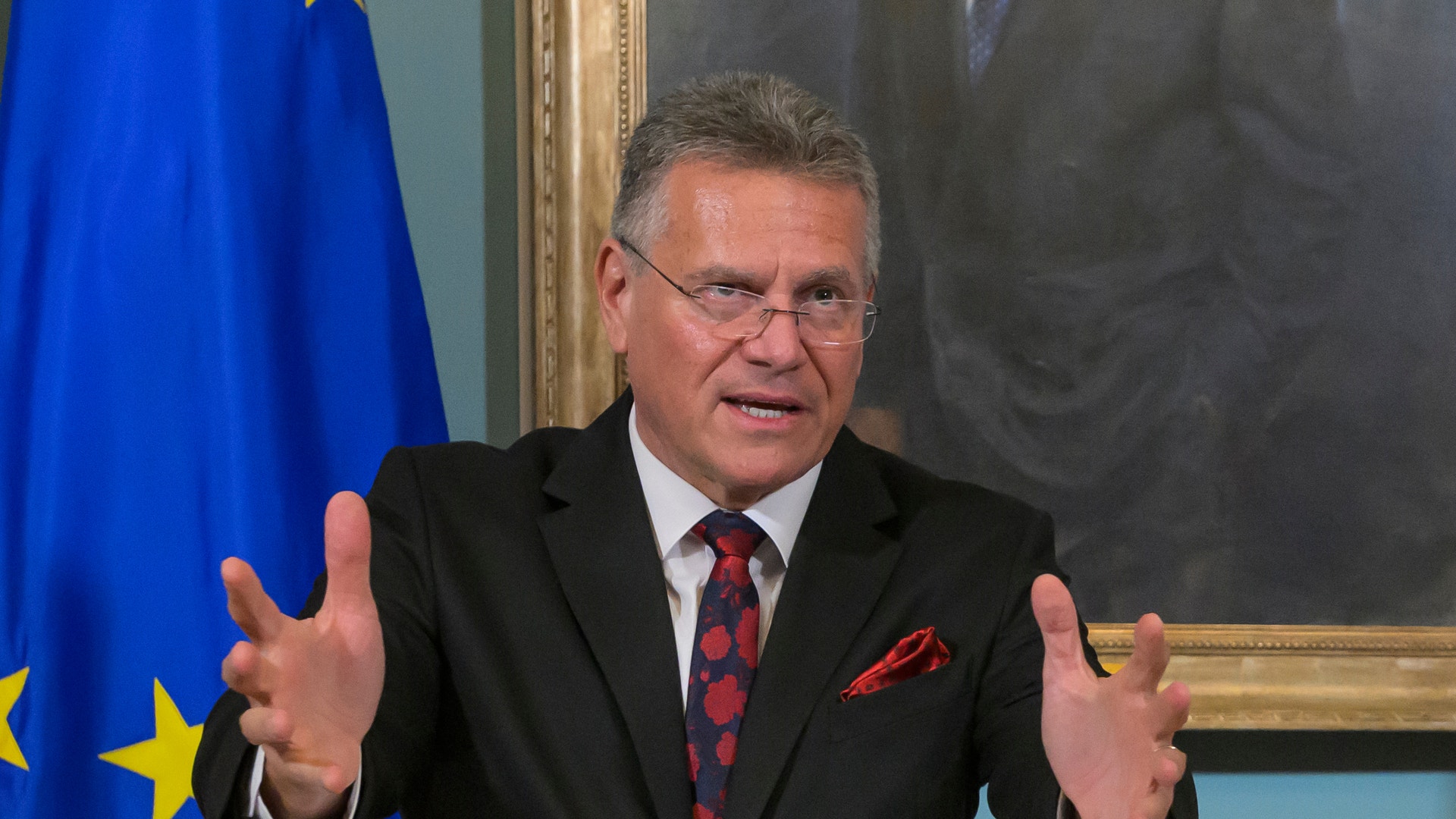 En man i svart kostym och röd slips talar vid ett podium. Han gestikulerar med båda händerna. I bakgrunden syns EU-flaggan och ett porträtt i guldram.
