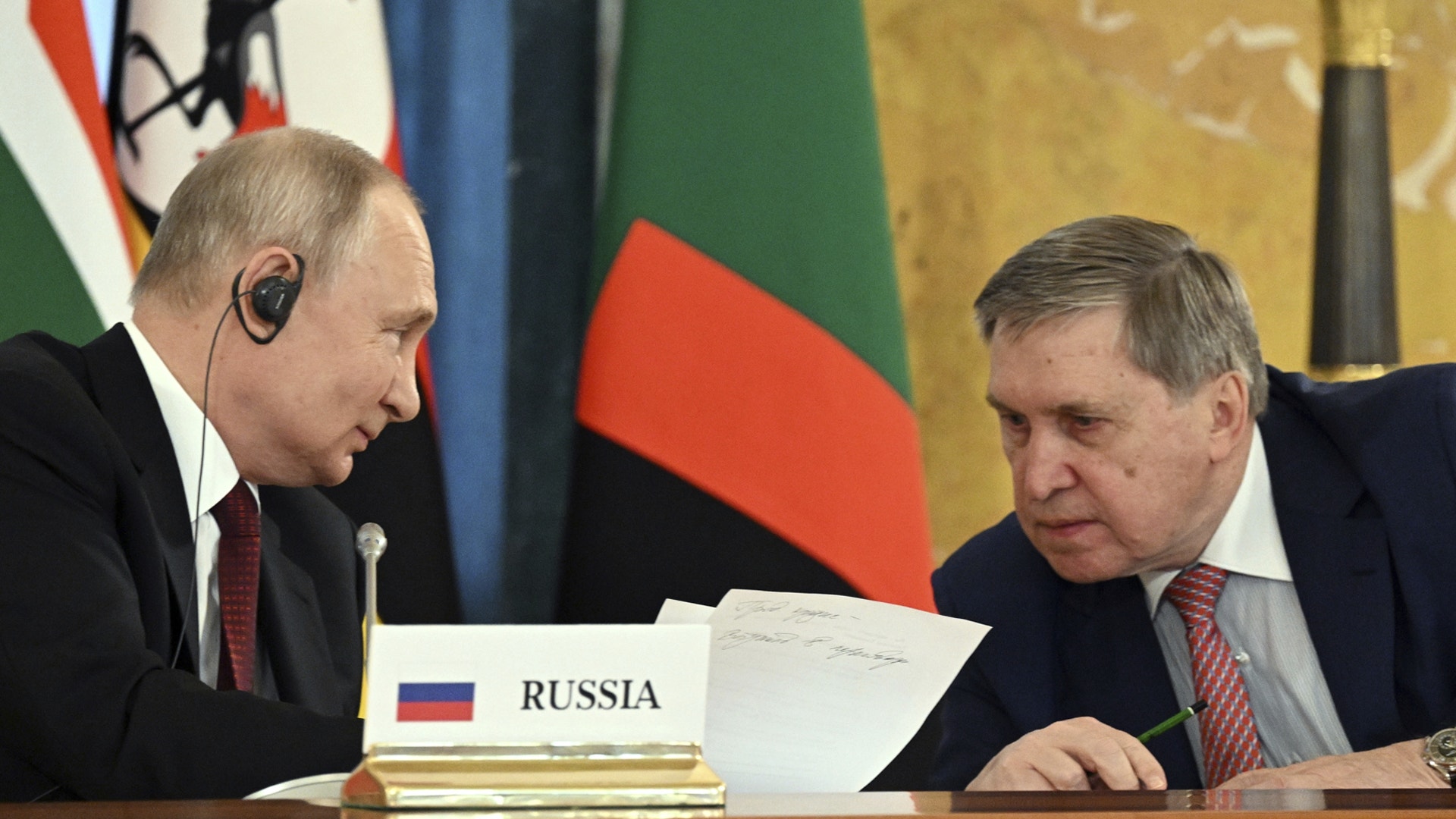 Två män i formell klädsel sitter vid ett mötesbord med flaggor i bakgrunden under en officiell sammankomst.