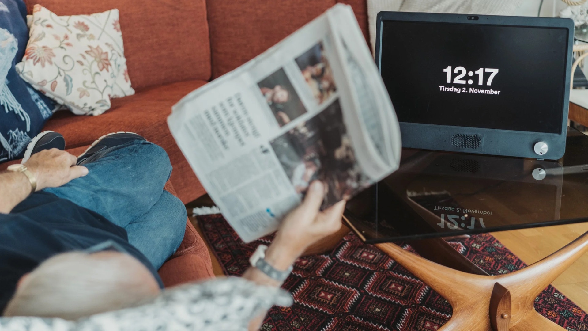 En person sitter på en röd soffa och läser en tidning. En tablet visar tiden 12:17 på bordet bredvid.