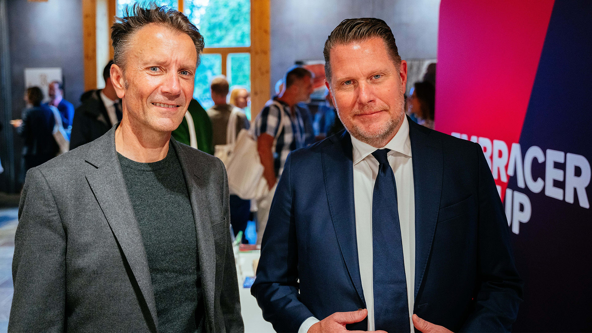 Två män står tillsammans på ett event. Mannen till vänster bär grå blazer och mörkgrön tröja. Mannen till höger bär mörkblå kostym med slips. I bakgrunden syns andra människor och en rosa vägg.