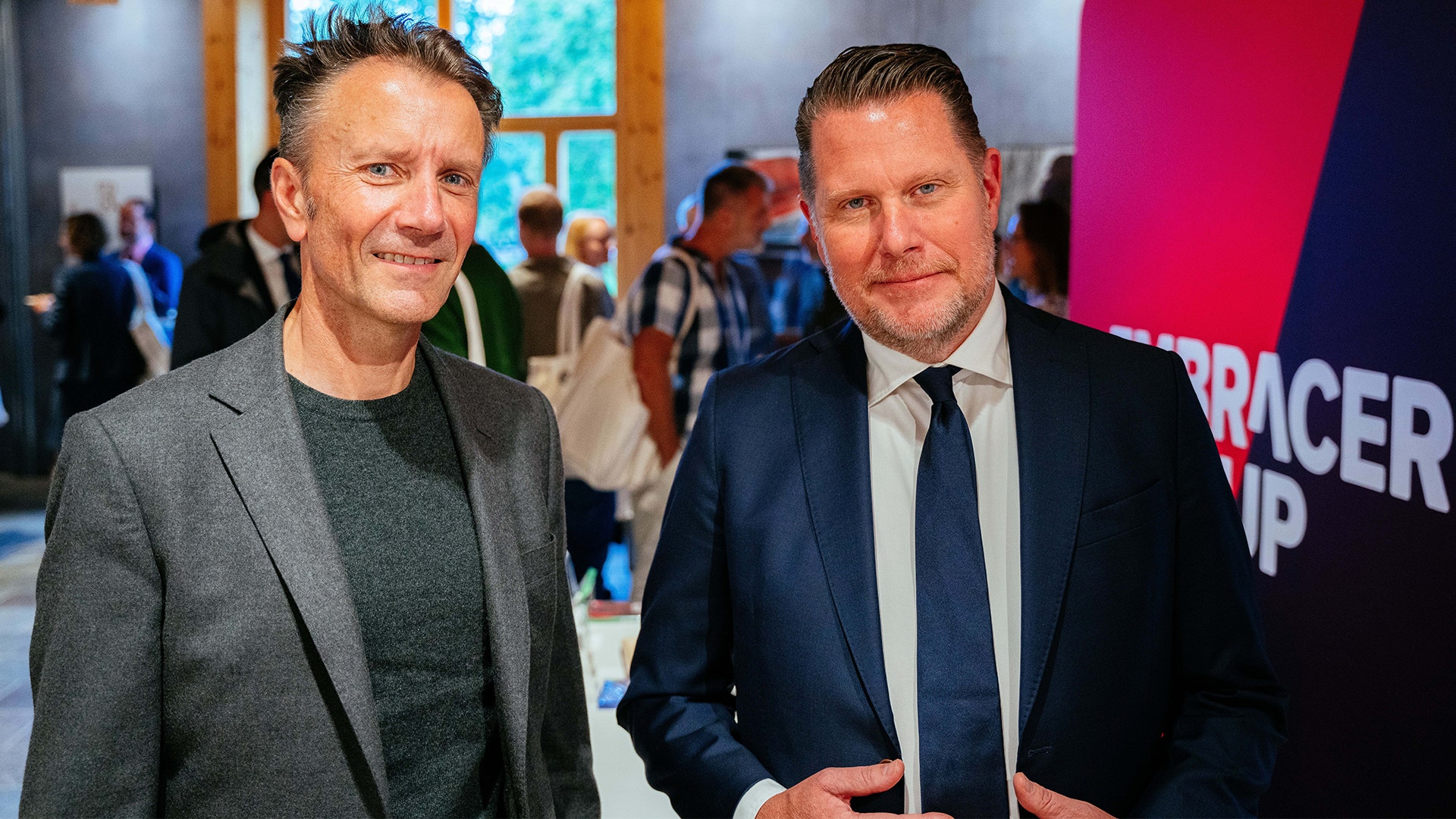 Två män står tillsammans på ett event. Mannen till vänster bär grå blazer och mörkgrön tröja. Mannen till höger bär mörkblå kostym med slips. I bakgrunden syns andra människor och en rosa vägg.