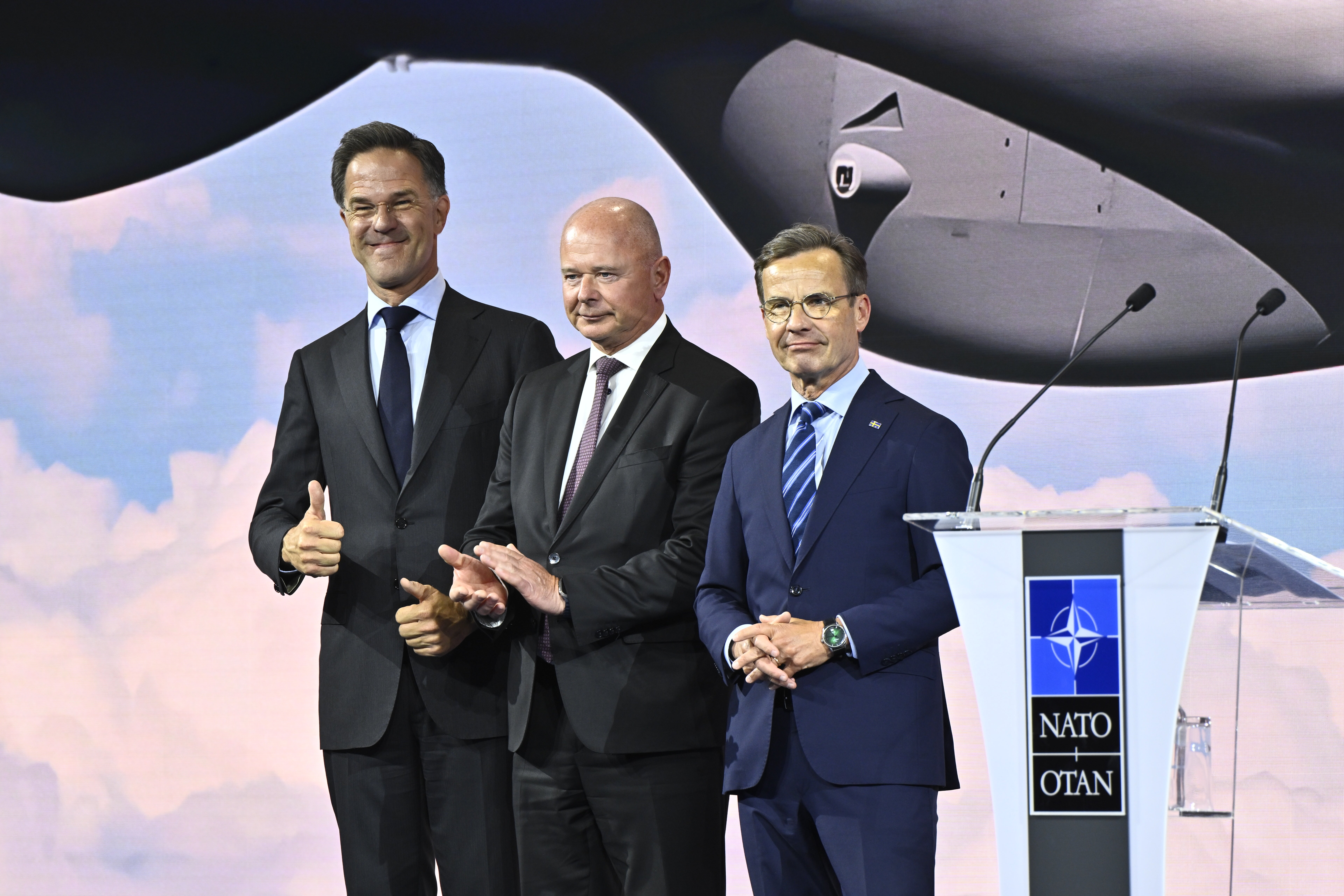 Tre män i mörka kostymer står vid ett NATO-podium framför ett militärflygplan.