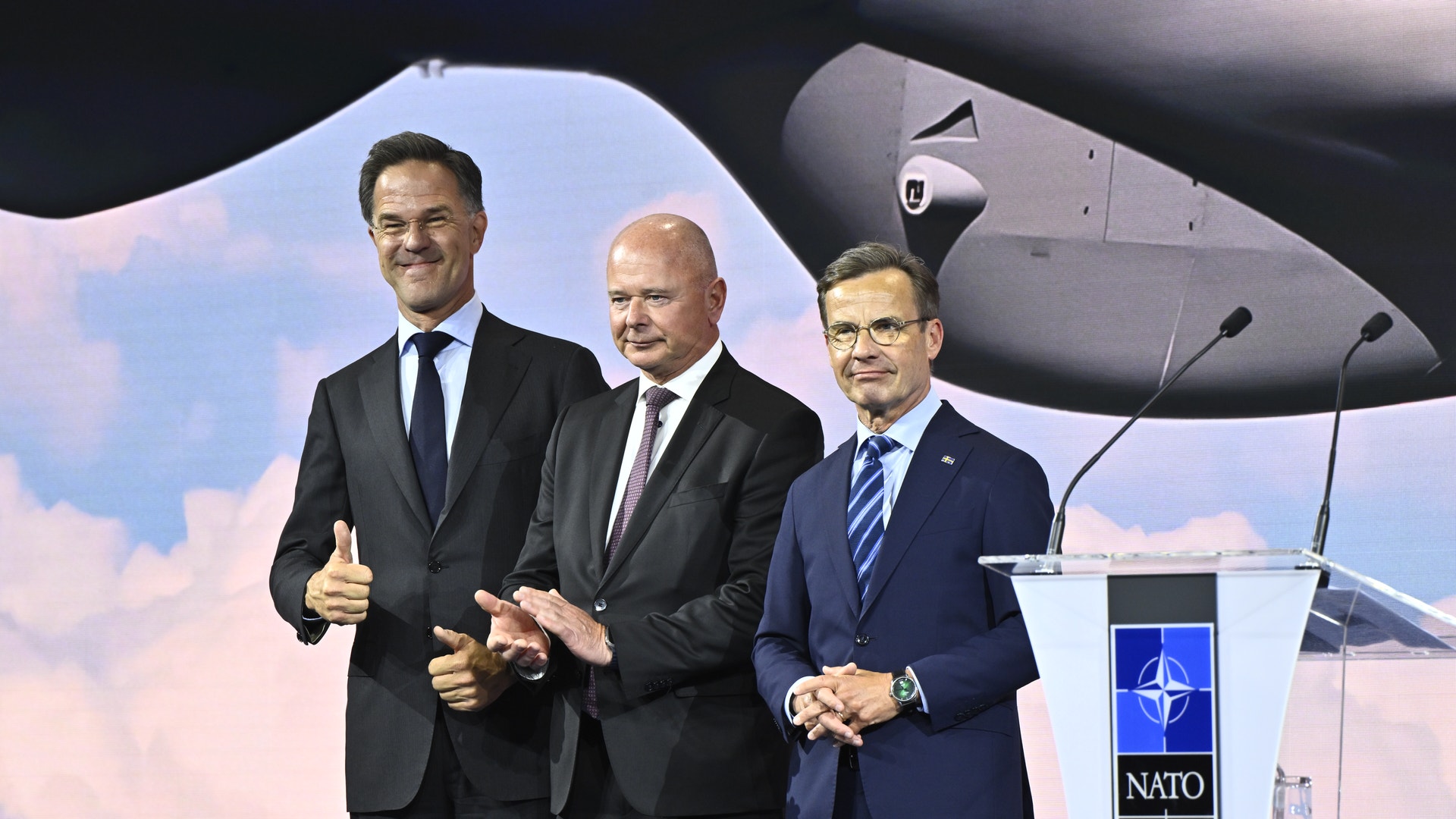 Tre män i mörka kostymer står vid ett NATO-podium framför ett militärflygplan.