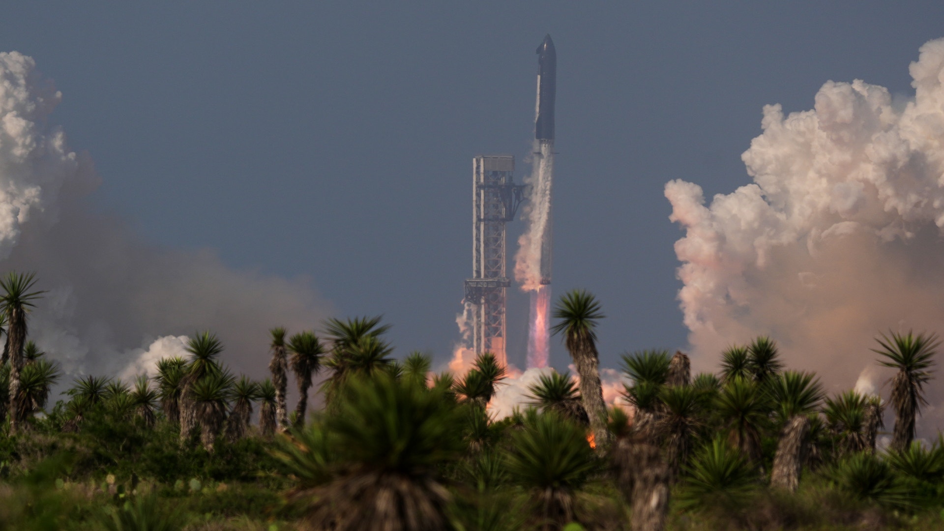 SpaceX Falcon Heavy-raket lyfter från startplatsen med aktiv förbränning. Palmer och vita moln syns i förgrunden och bakgrunden.