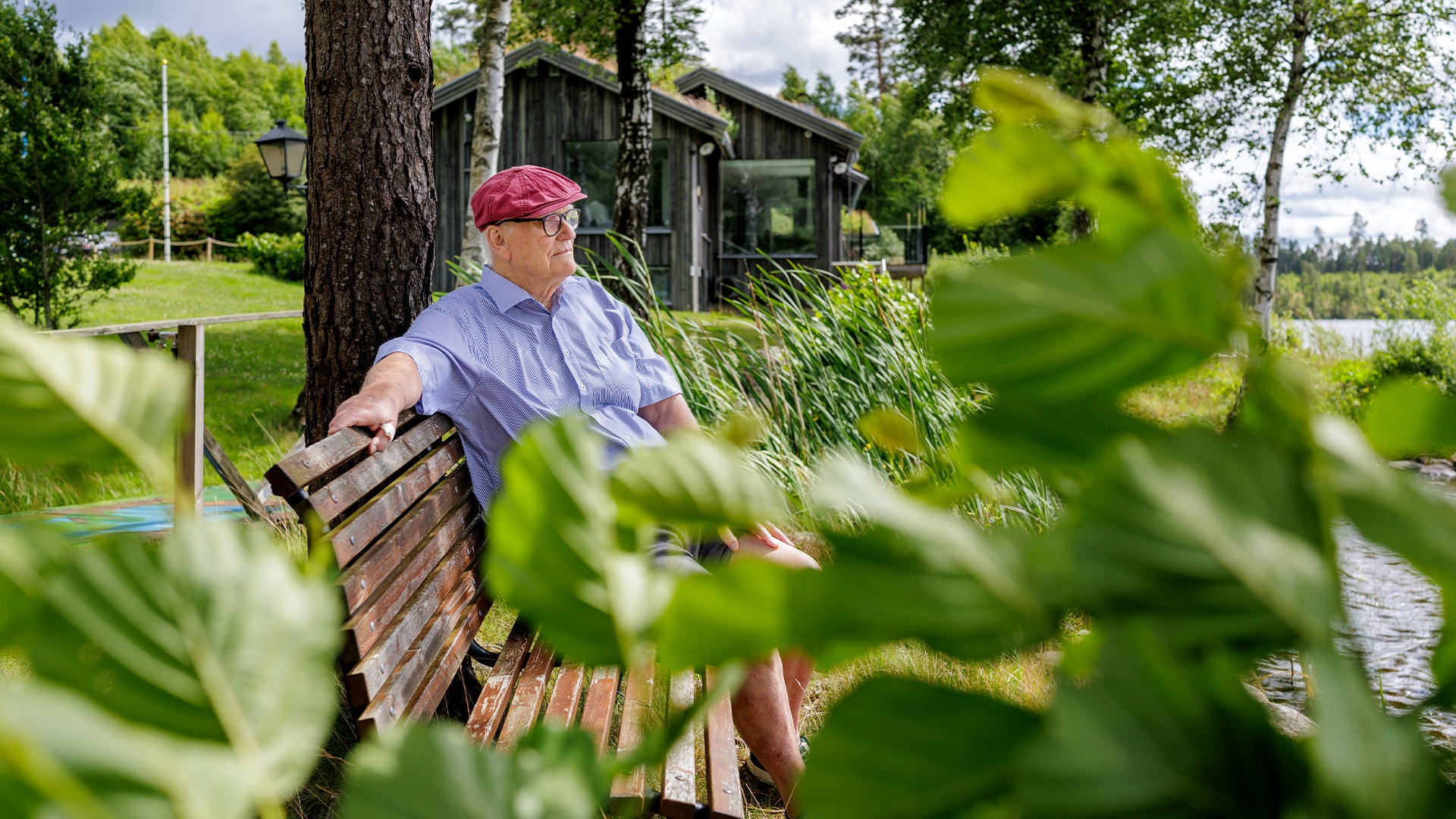 En man i ljusblå skjorta och röd keps sitter på en träbänk vid vattnet, omgiven av gröna växter och träd.