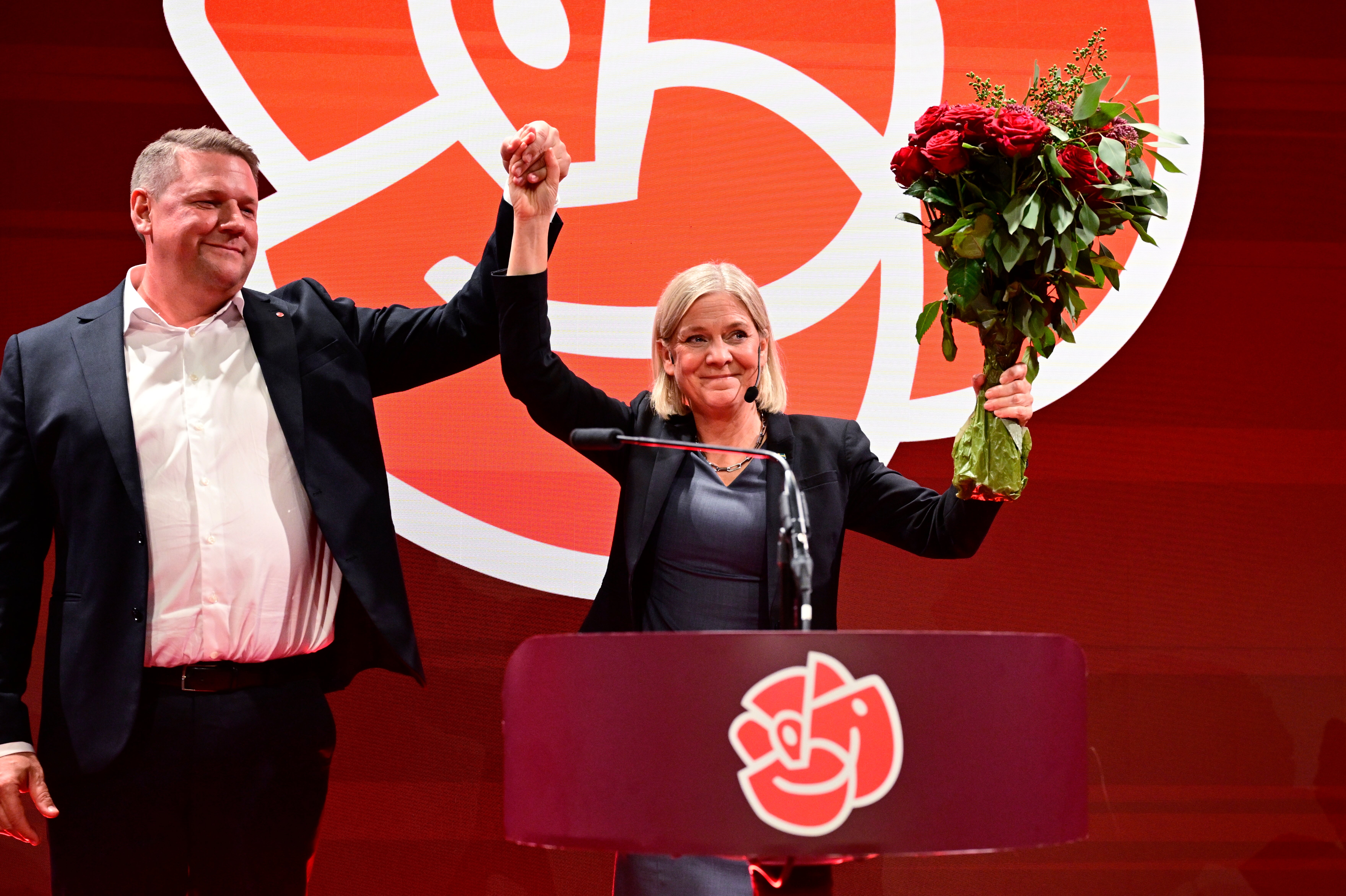 En man och en kvinna på en scen håller upp varandras händer i luften, kvinnan håller en bukett rosor, i bakgrunden syns Socialdemokraternas logotyp.