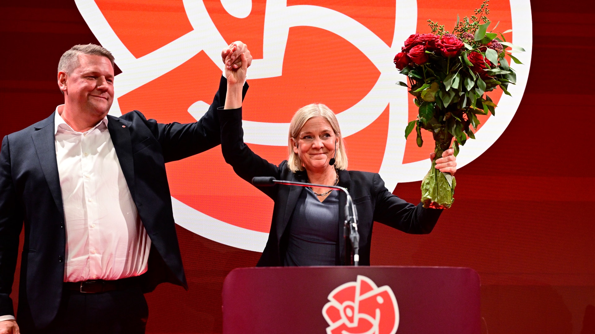 En man och en kvinna på en scen håller upp varandras händer i luften, kvinnan håller en bukett rosor, i bakgrunden syns Socialdemokraternas logotyp.