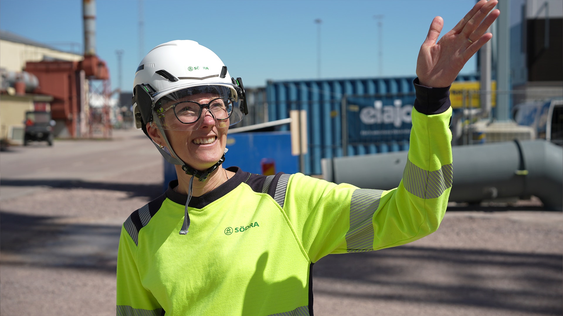 En person i gula reflekterande arbetskläder och vit säkerhetshjalm vinkar på en industriell arbetsplats med containrar och industriella installationer i bakgrunden.