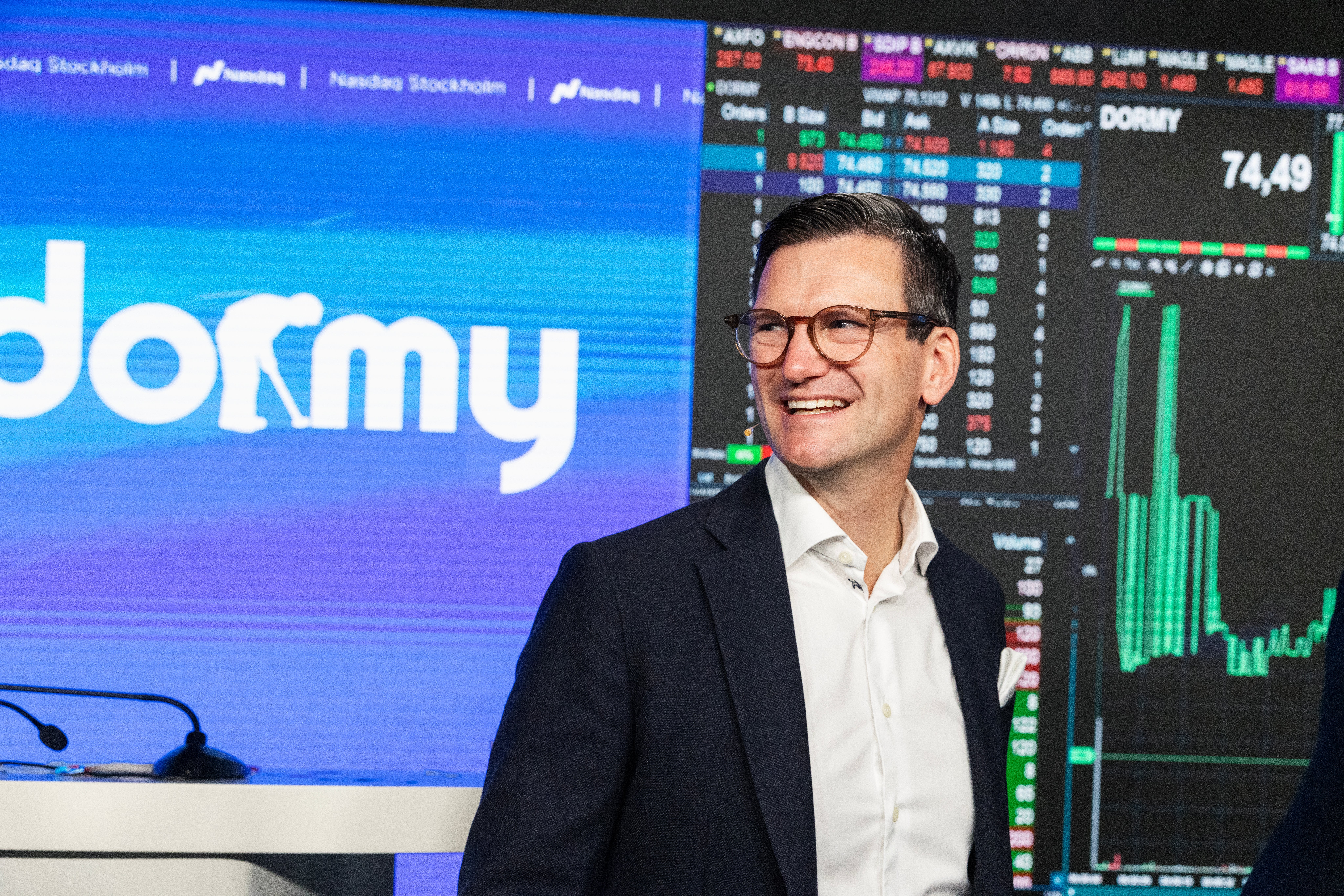 En leende man i kostym står framför storbildsskärmar med Dormys logotyp och börskurser i samband med bolagets notering på Nasdaq Stockholm.