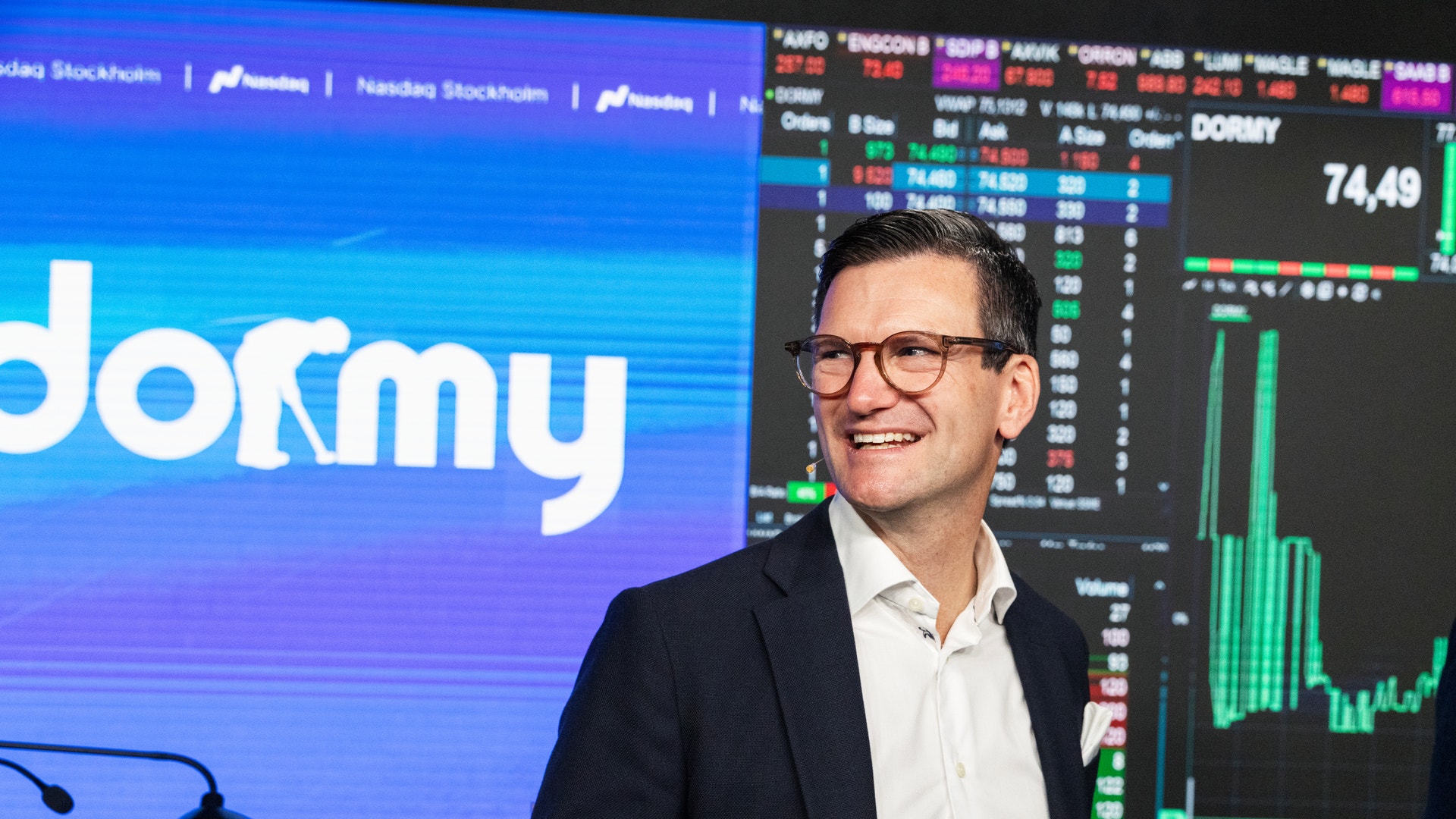En leende man i kostym står framför storbildsskärmar med Dormys logotyp och börskurser i samband med bolagets notering på Nasdaq Stockholm.