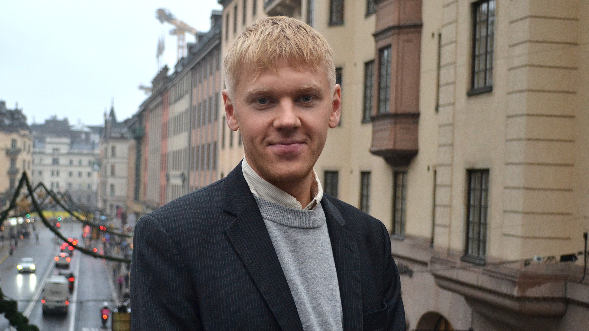 Porträtt av en man med blont hår, klädd i mörk kavaj och grå polotröja, fotograferad framför klassiska europeiska byggnader.