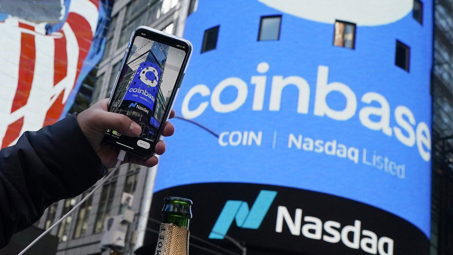 En person håller upp en smartphone med Coinbase-logotypen framför en stor blå väggskärm med Coinbase och Nasdaq-logotyper.